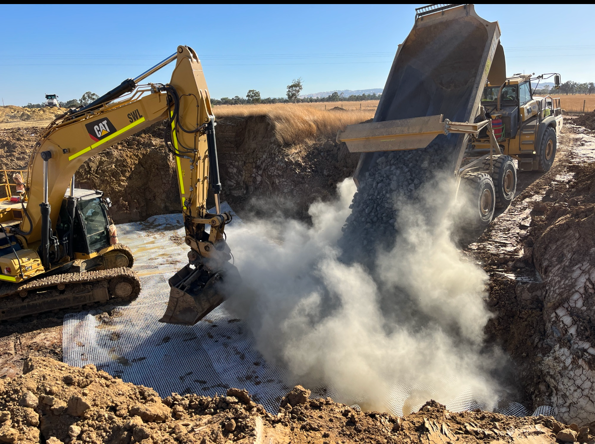 An excavator and a dump truck are working together at a construction site, with the dump truck pouring dirt into the excavator's bucket as it digs into the ground.