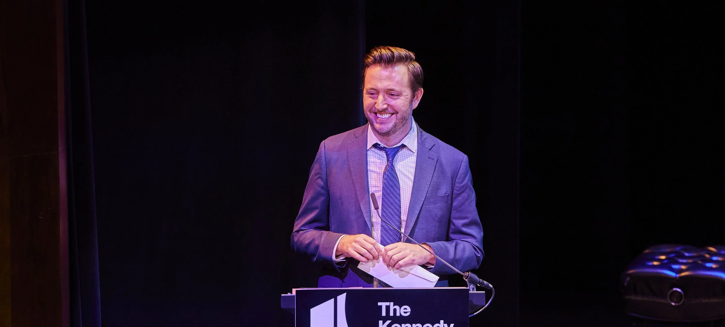 A man wearing a gray suit, light blue shirt, and blue tie stands at a podium with a microphone, smiling and holding a piece of paper. The podium has a sign that says "The Kennedy" against a dark background.