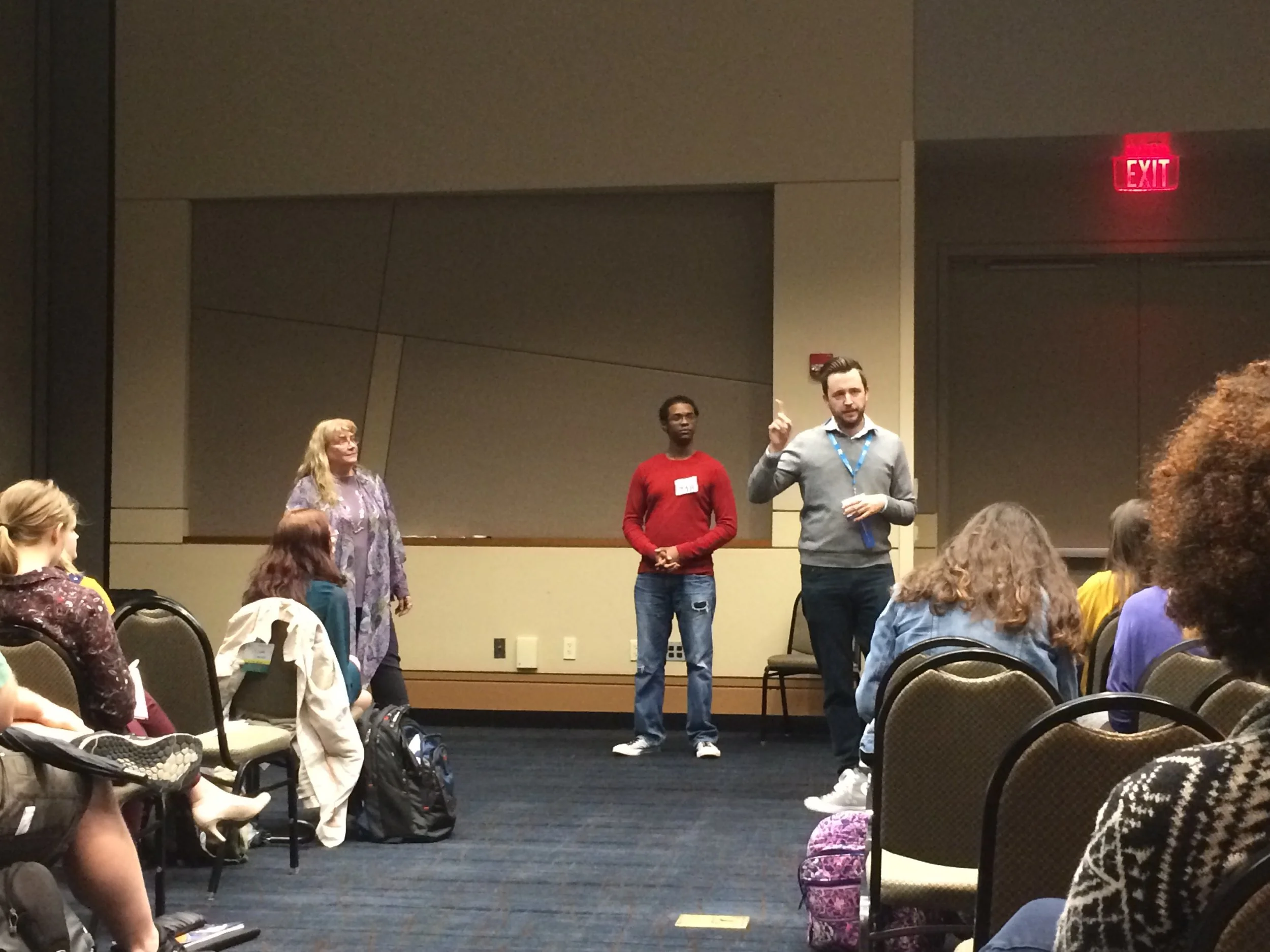 A group of people is participating in a presentation or discussion in a conference room with chairs. Three individuals are standing at the front, one woman and two men, speaking to an audience of seated participants.