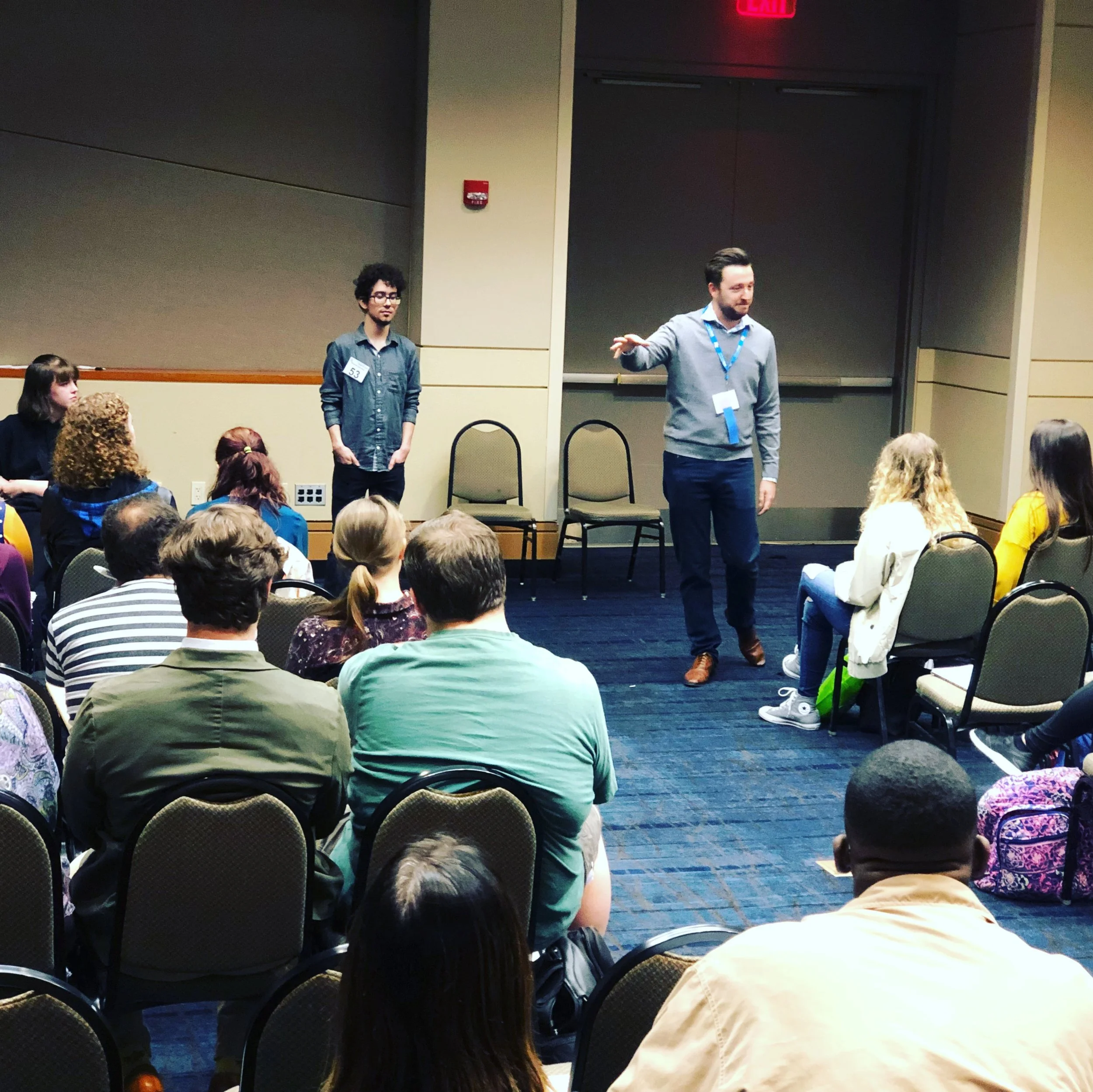 A man with short dark hair and a beard, wearing a gray sweater and a blue lanyard, is speaking to a group in a conference room. Next to him, a young man with curly dark hair and glasses, dressed in a dark shirt, stands with a badge on his chest. The audience, consisting of diverse people, is seated and listening attentively.