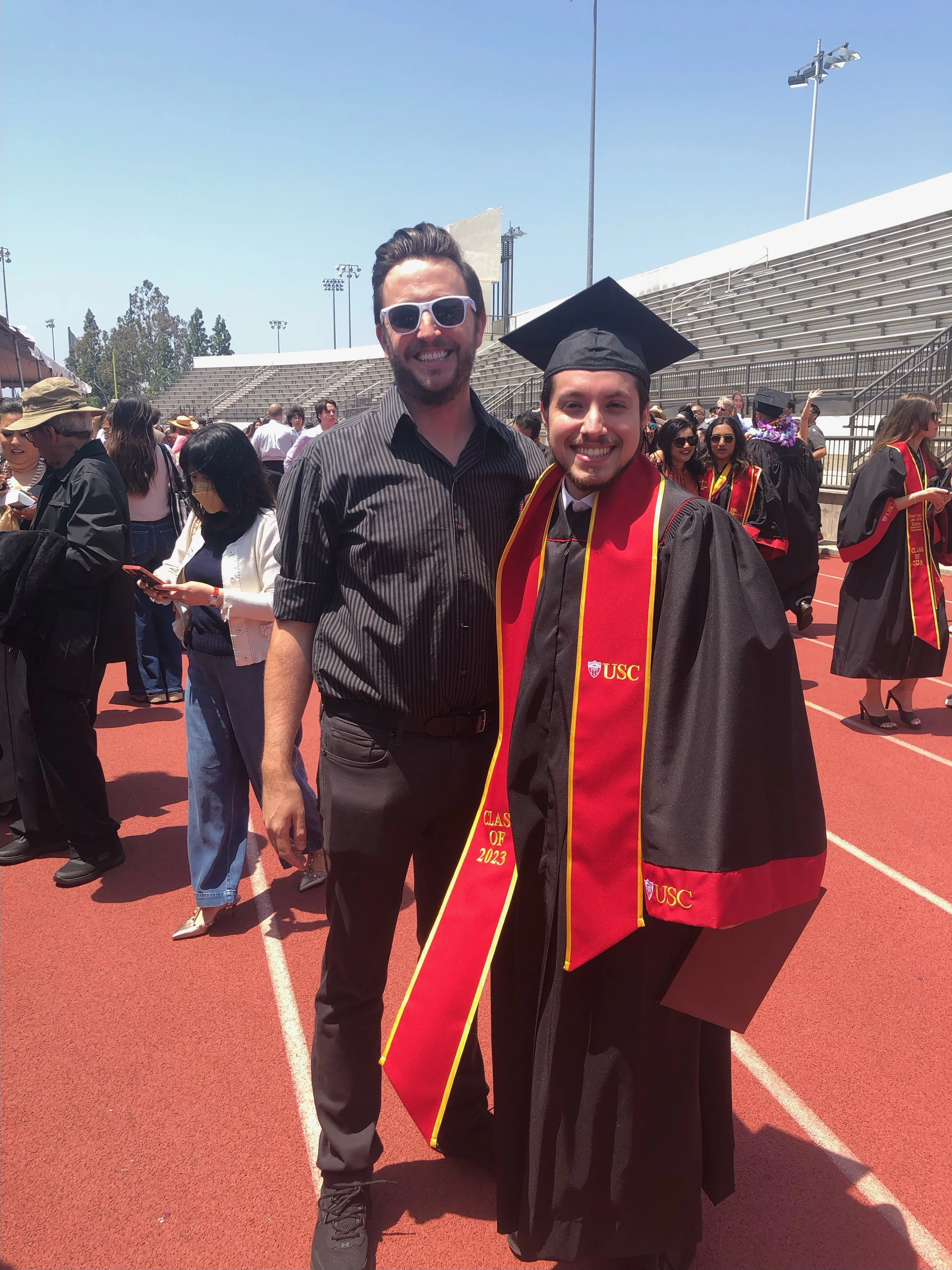 A man and a graduate wearing a black cap and gown with a red and yellow sash at an outdoor graduation ceremony at USC on a sunny day. The man is smiling and wearing sunglasses, and the graduate has a big smile.