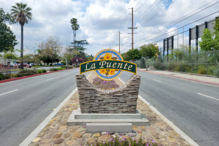 A street sign for La Puente in a landscaped median with rocks and flowers, set against a background of trees, parked cars, utility poles, and a cloudy sky.