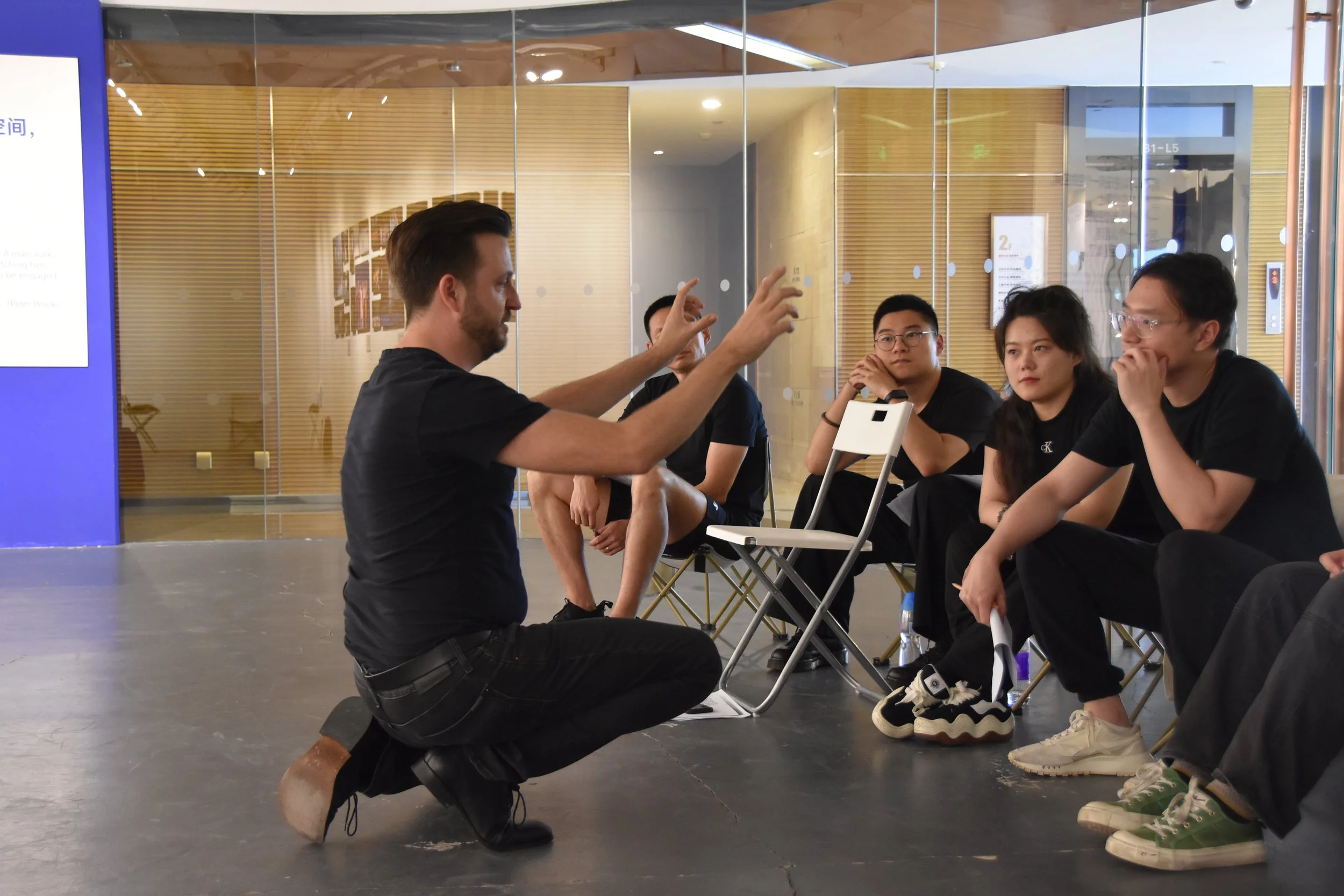 A man kneeling and speaking to four seated young adults in a modern indoor space with wooden wall panels and glass walls.