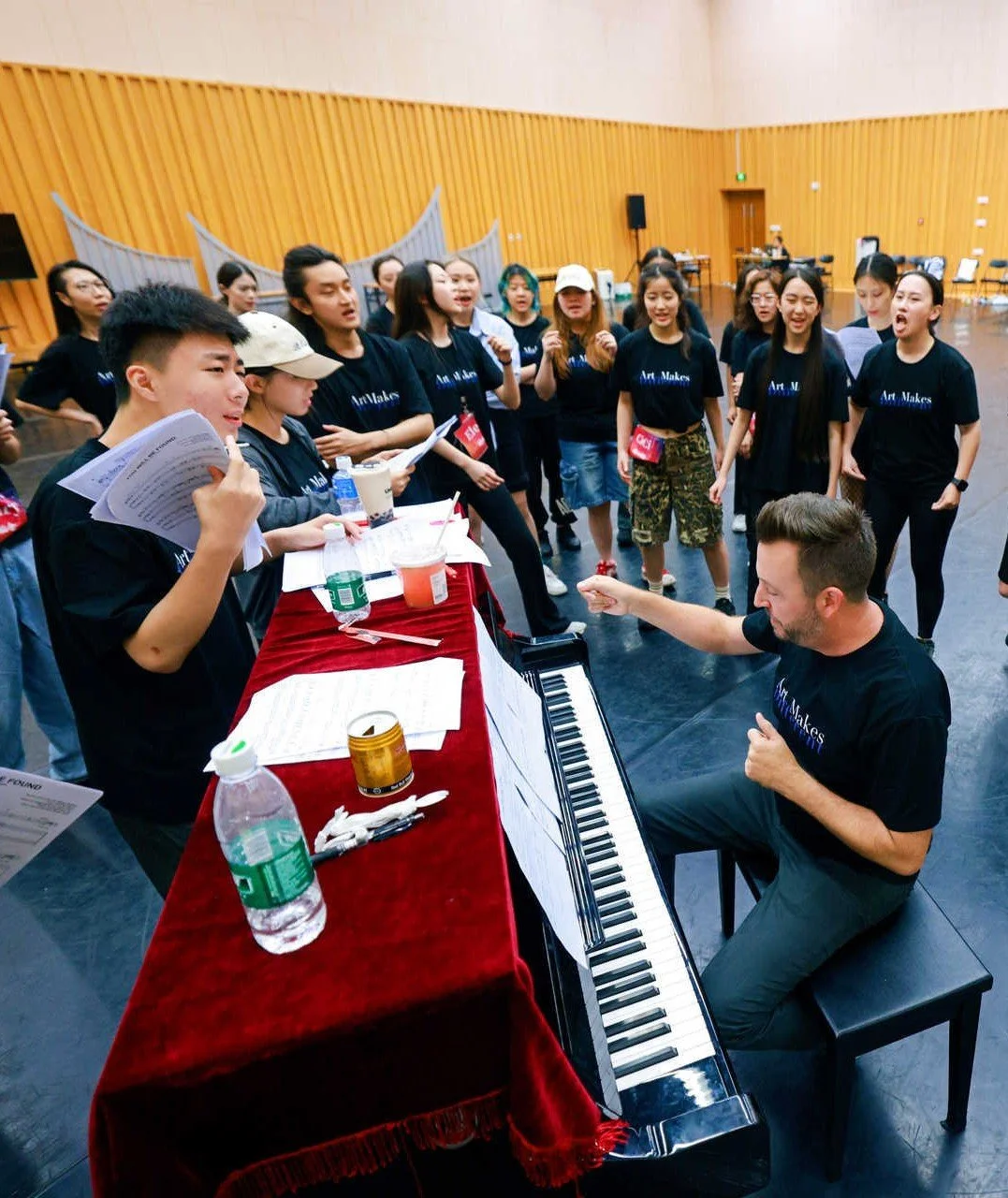 A group of young people gathers around a man playing the piano in a large rehearsal space or auditorium. The group appears to be singing along, holding sheet music, with some smiling and engaging actively. The man at the piano is pointing and conducting confidently. The room has wooden paneling and folding chairs in the background.