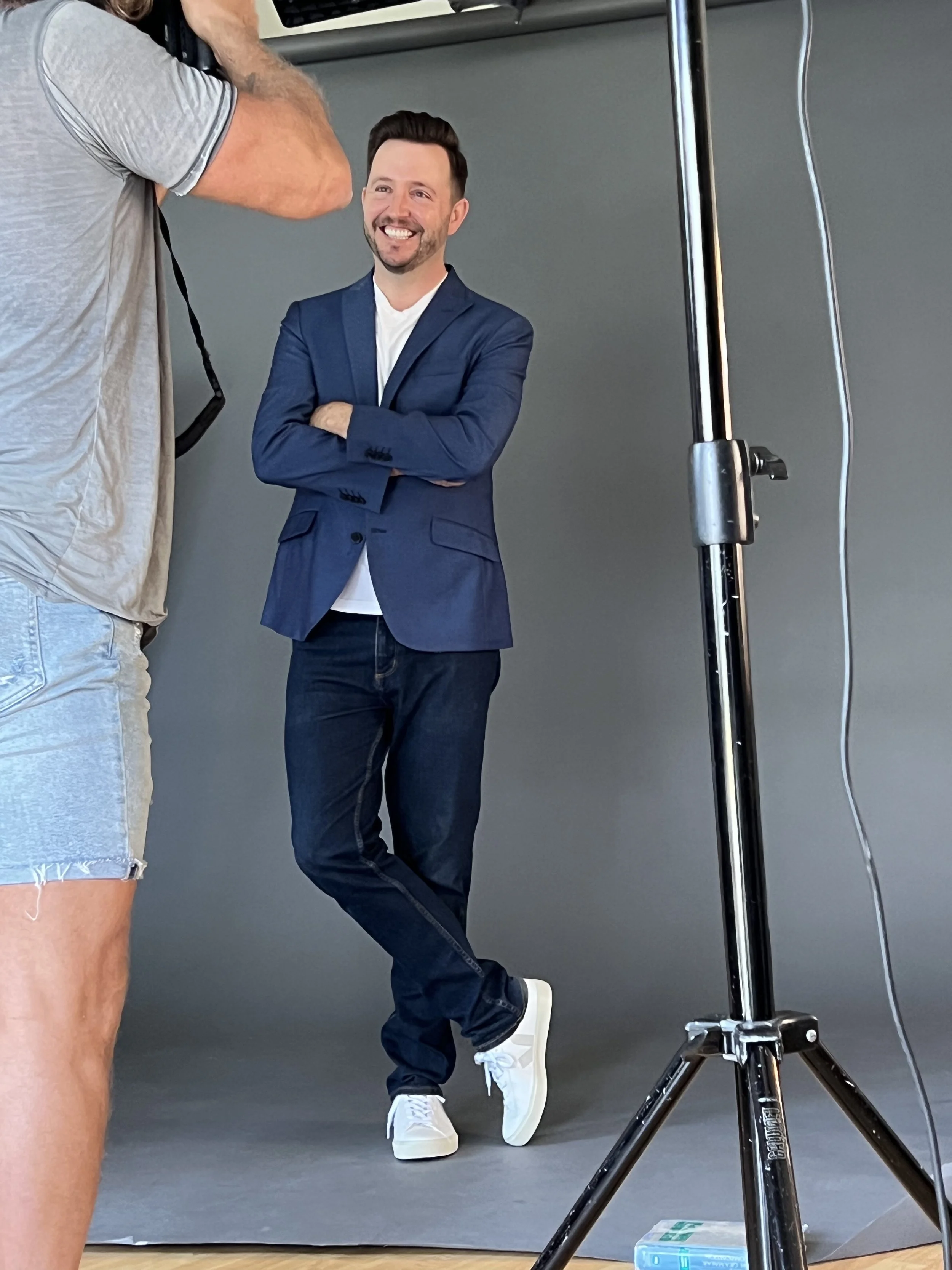 A man with dark hair and a beard smiling and crossing his arms, dressed in a blue blazer and jeans, standing in front of a plain grey background during a photoshoot, with a photography light stand visible on the right.