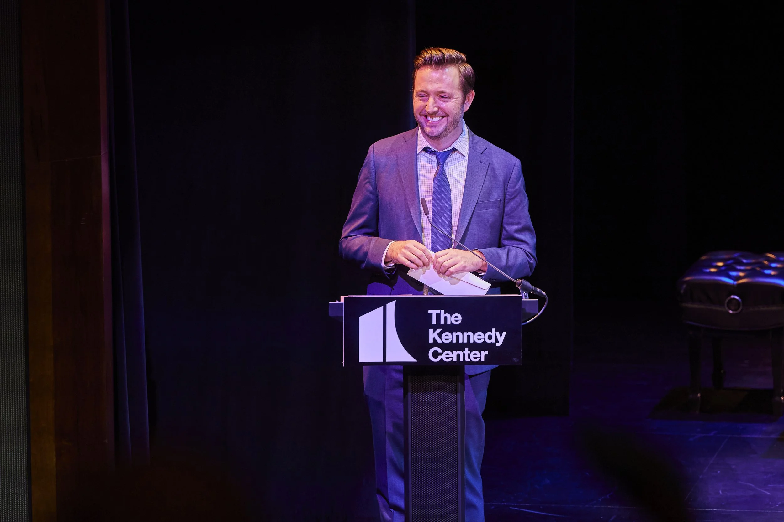 A man in a suit stands smiling behind a podium at The Kennedy Center.