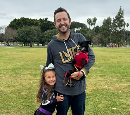 A smiling man holding a black dog in a red sweater in a park, with a young girl standing beside him smiling.