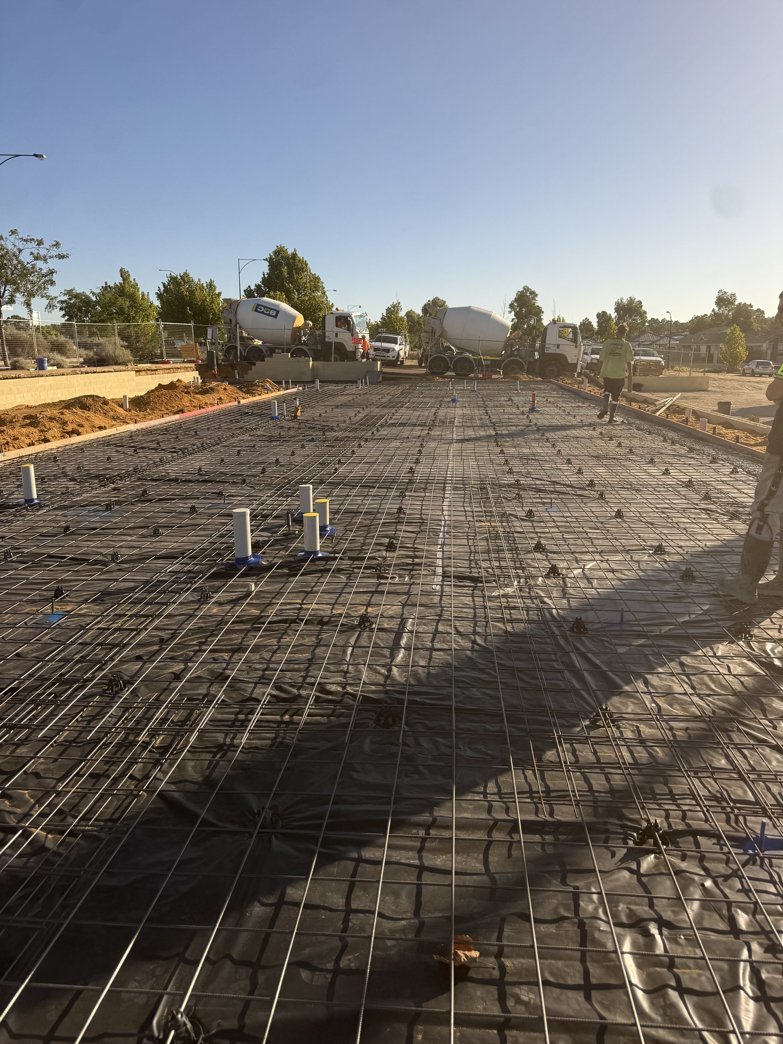 Construction site with steel rebar laid out for a concrete slab, workers are present, and cement mixers are visible in the background during daytime.