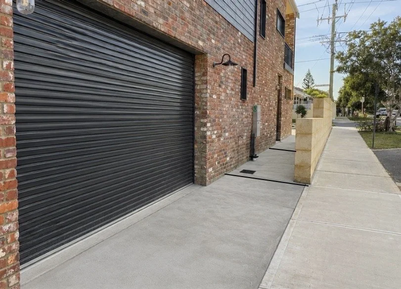 Sidewalk and driveway outside modern brick building with garage door, small windows, and a brick and stone wall on sidewalk.