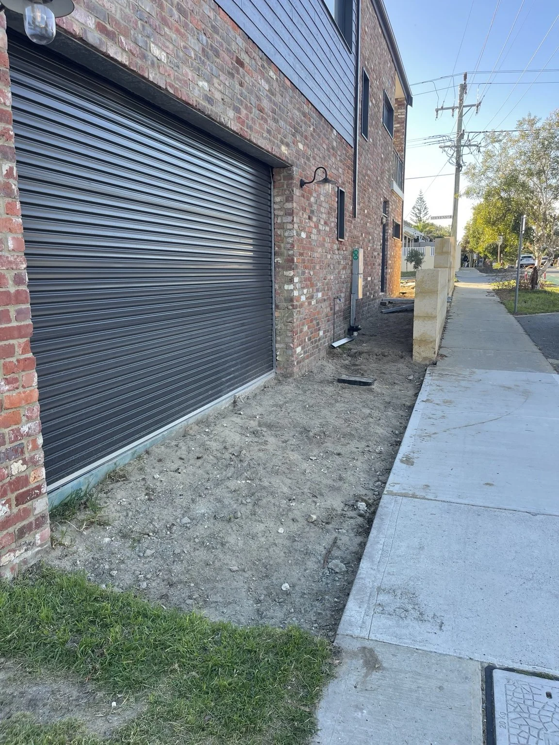 Sidewalk next to a brick building with a black metal garage door. Soil and some grass are visible near the base of the building. There are some construction materials and a small wall on the sidewalk. Utility poles and trees are in the background.