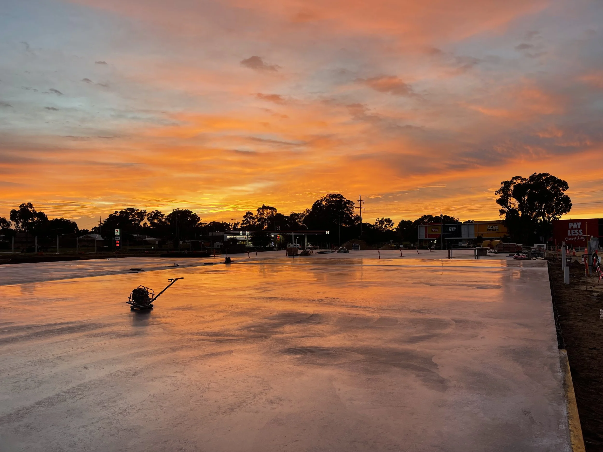 A sunset sky with orange, pink, and purple clouds over a parking lot with trees and commercial buildings.