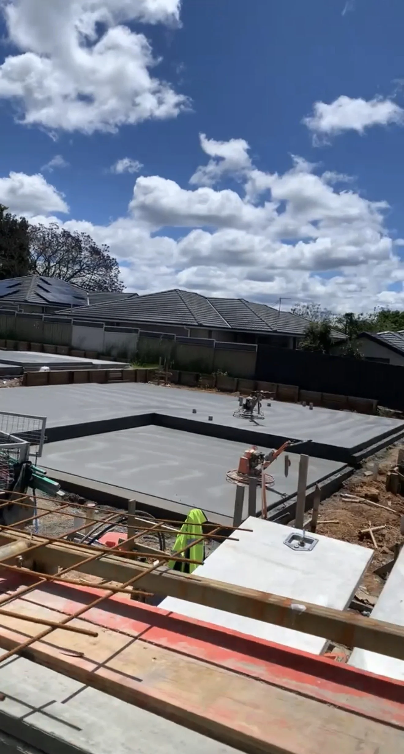 Construction site with a concrete foundation for a building, construction tools and equipment, and residential houses with tiled roofs in the background under a partly cloudy sky.