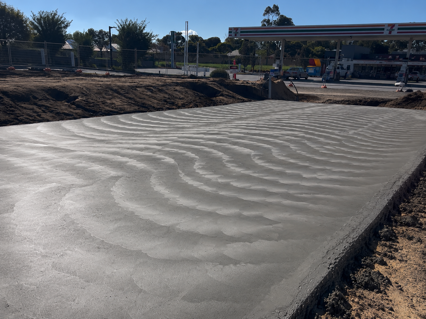 Freshly poured concrete slab with visible trowel marks at a construction site near a gas station, with fencing and trees in the background.