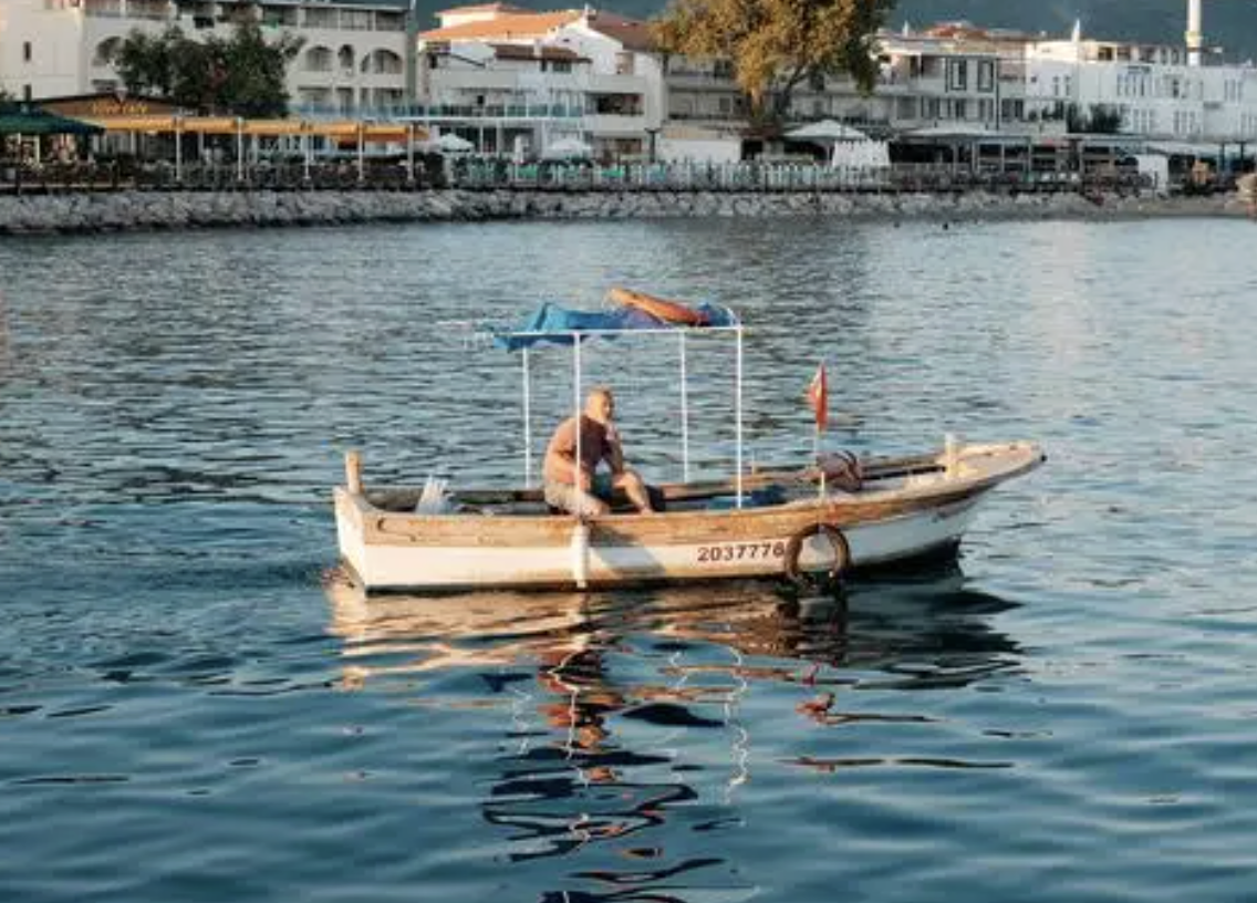 A person sitting on a small boat with a canopy on a calm body of water, with a shoreline lined with white buildings and trees in the background during daytime.