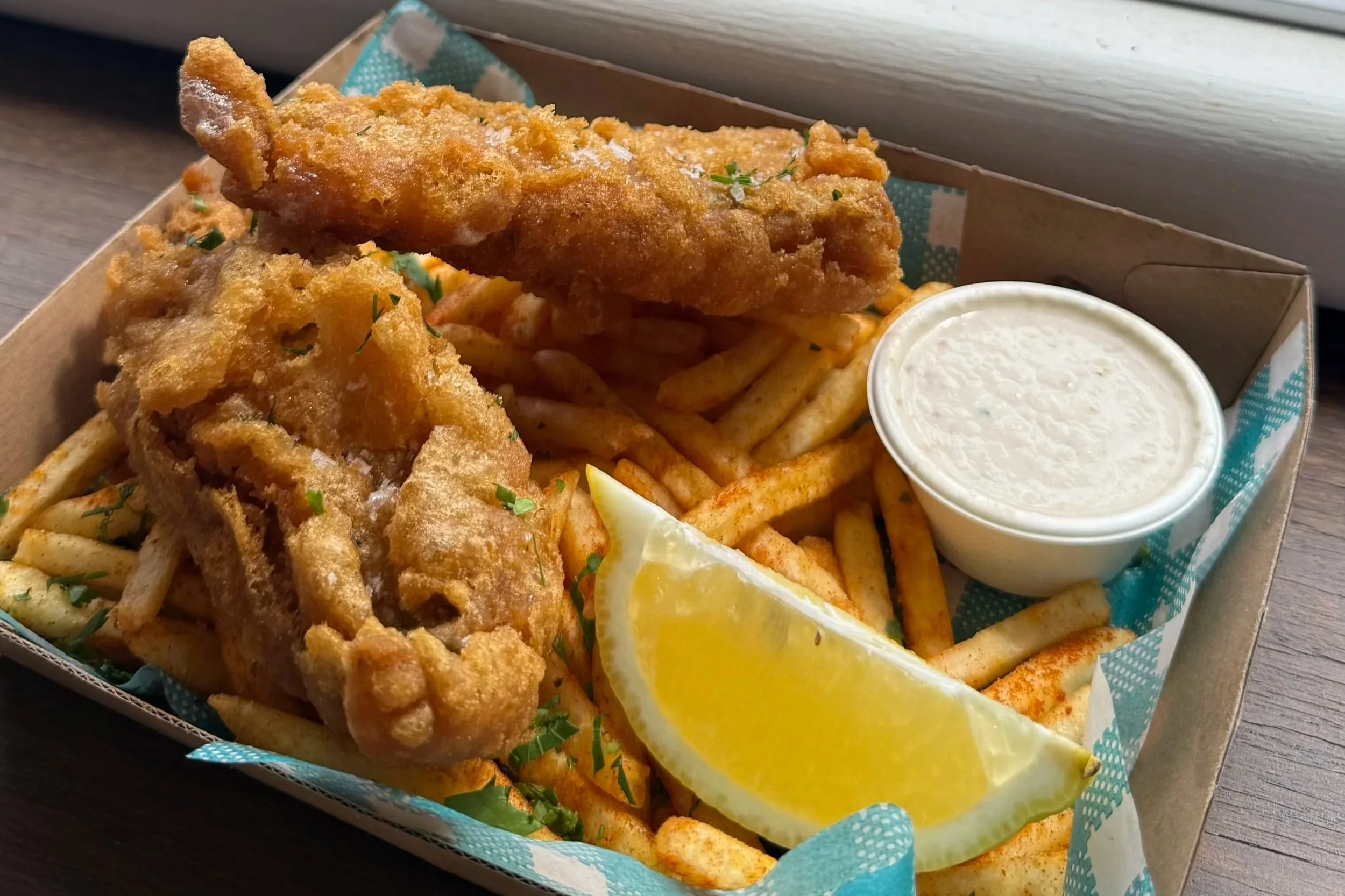 Fried fish with French fries, a slice of lemon, and a side of tartar sauce in a takeout box.
