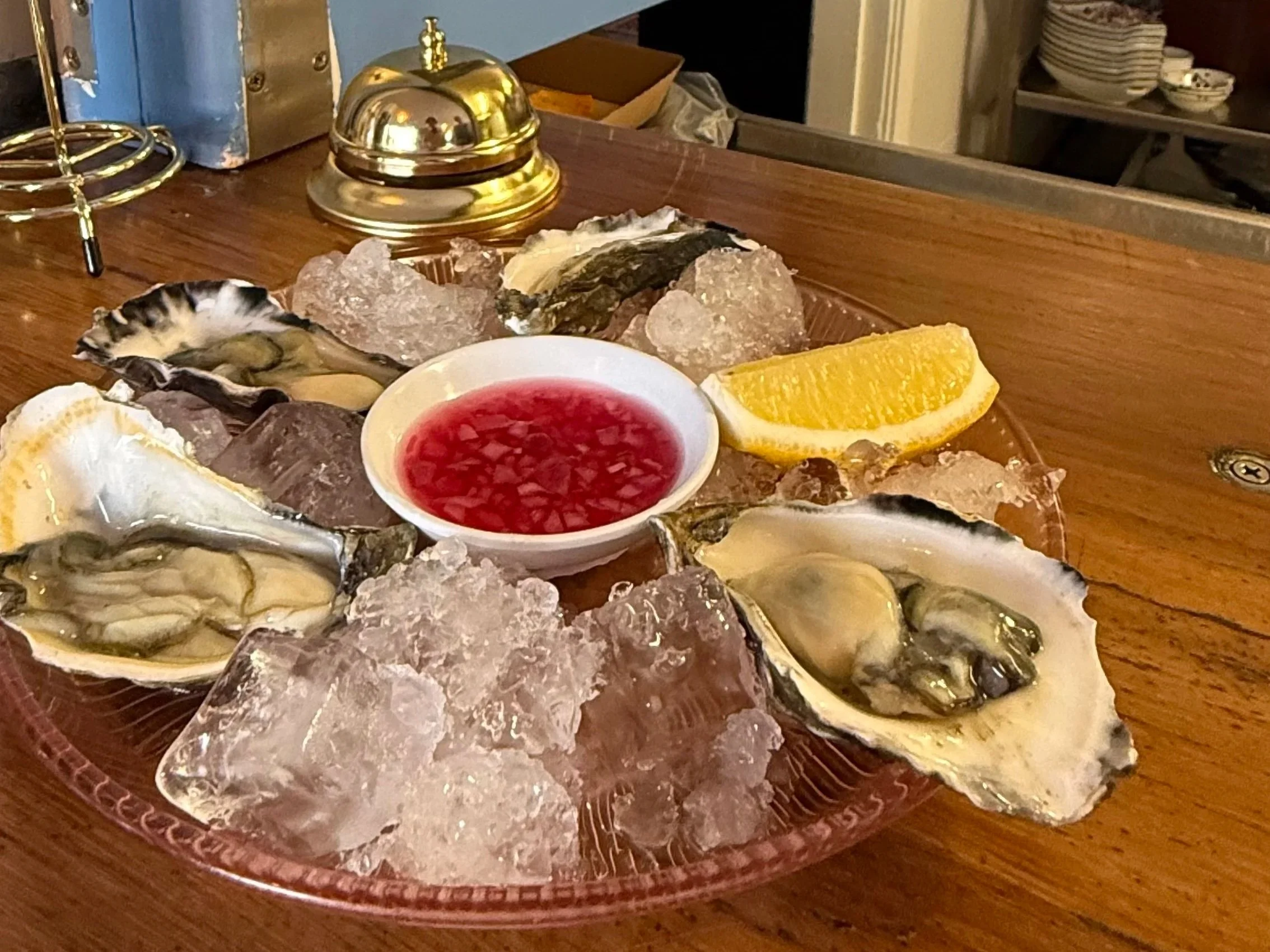 A round red tray holding fresh oysters on the half shell with Lemon wedge, cocktail sauce, ice and decorative gold-colored item on a wooden table.