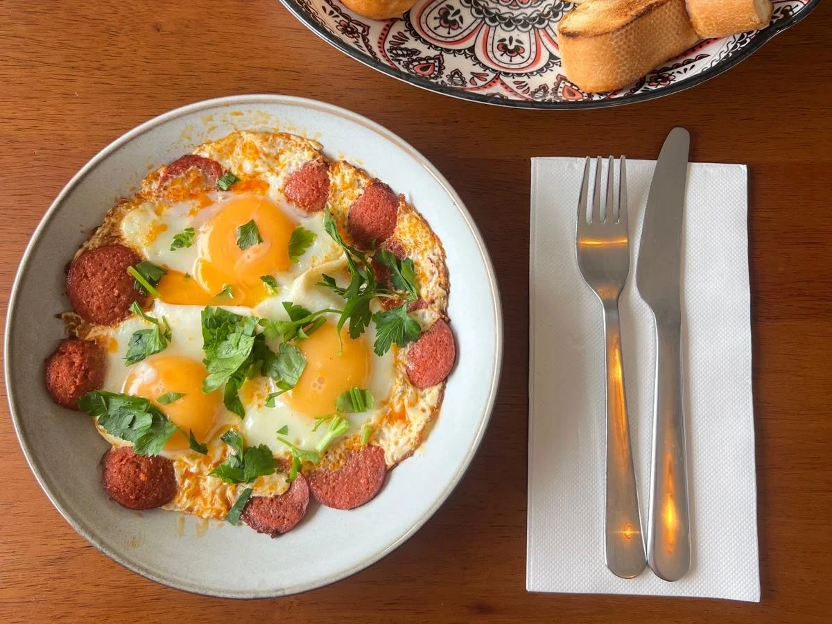 A plate with three fried eggs topped with parsley, surrounded by slices of sausage, on a wooden table. To the right, a fork and knife on a white napkin. In the upper right corner, there is a bowl of sliced baguette.