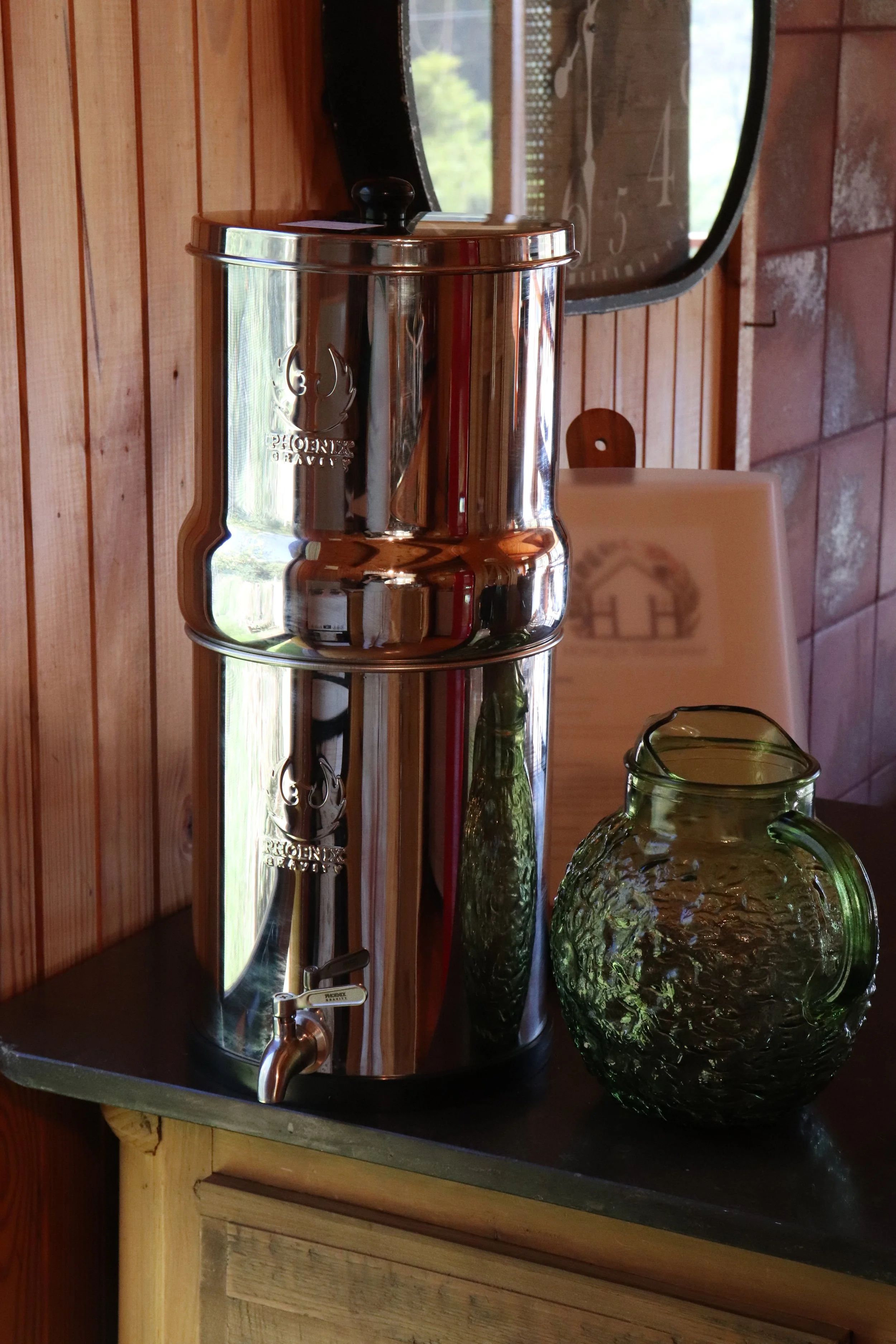 A large, shiny metal coffee urn with a spigot at the bottom, placed on a dark countertop in a rustic setting. There are two large glass vases with textured green surfaces nearby, on the right. A wooden wall with horizontal planks is behind the urn, a