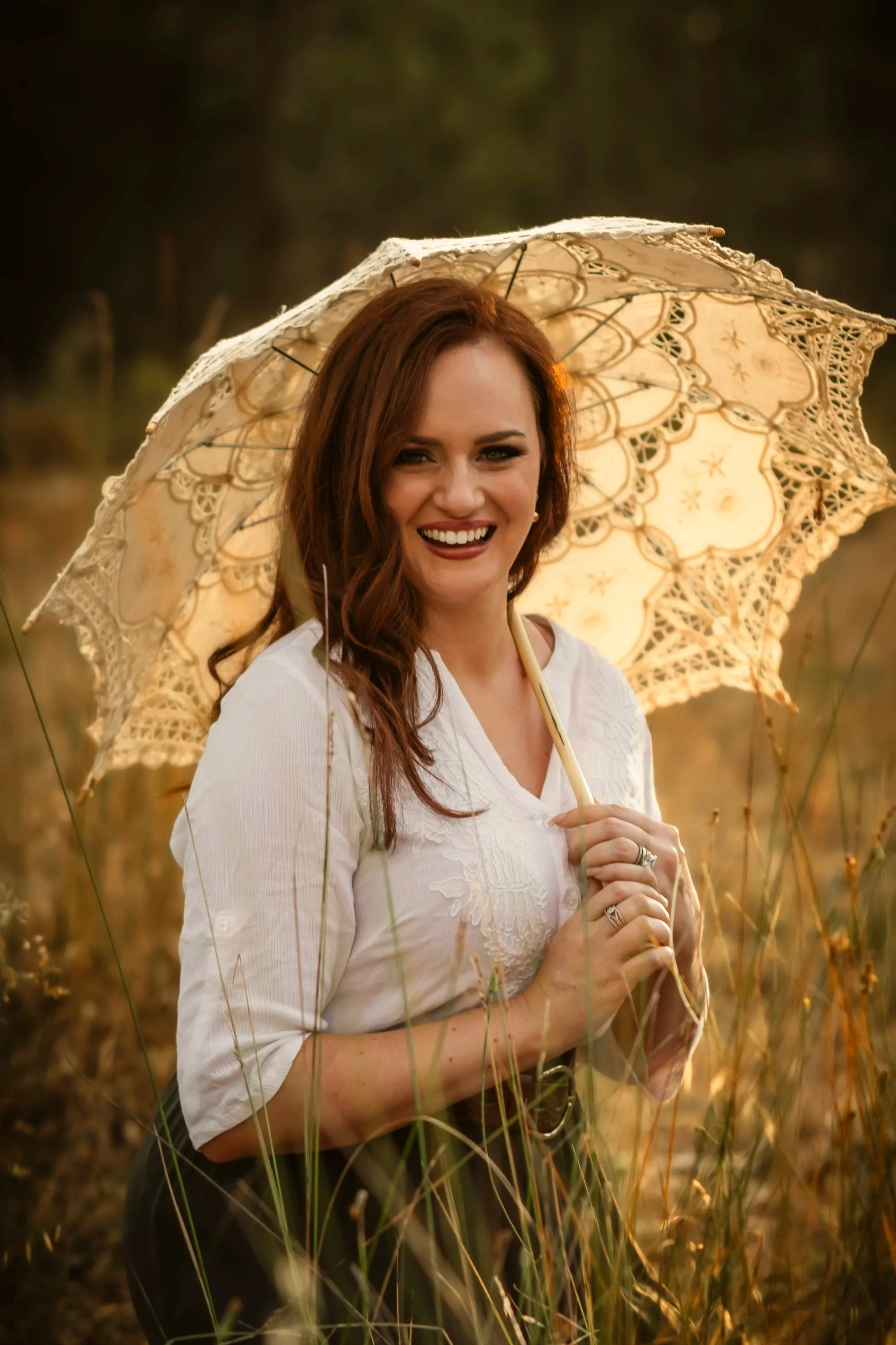 A woman with long red hair smiling while holding a decorative lace parasol in a field during sunset.