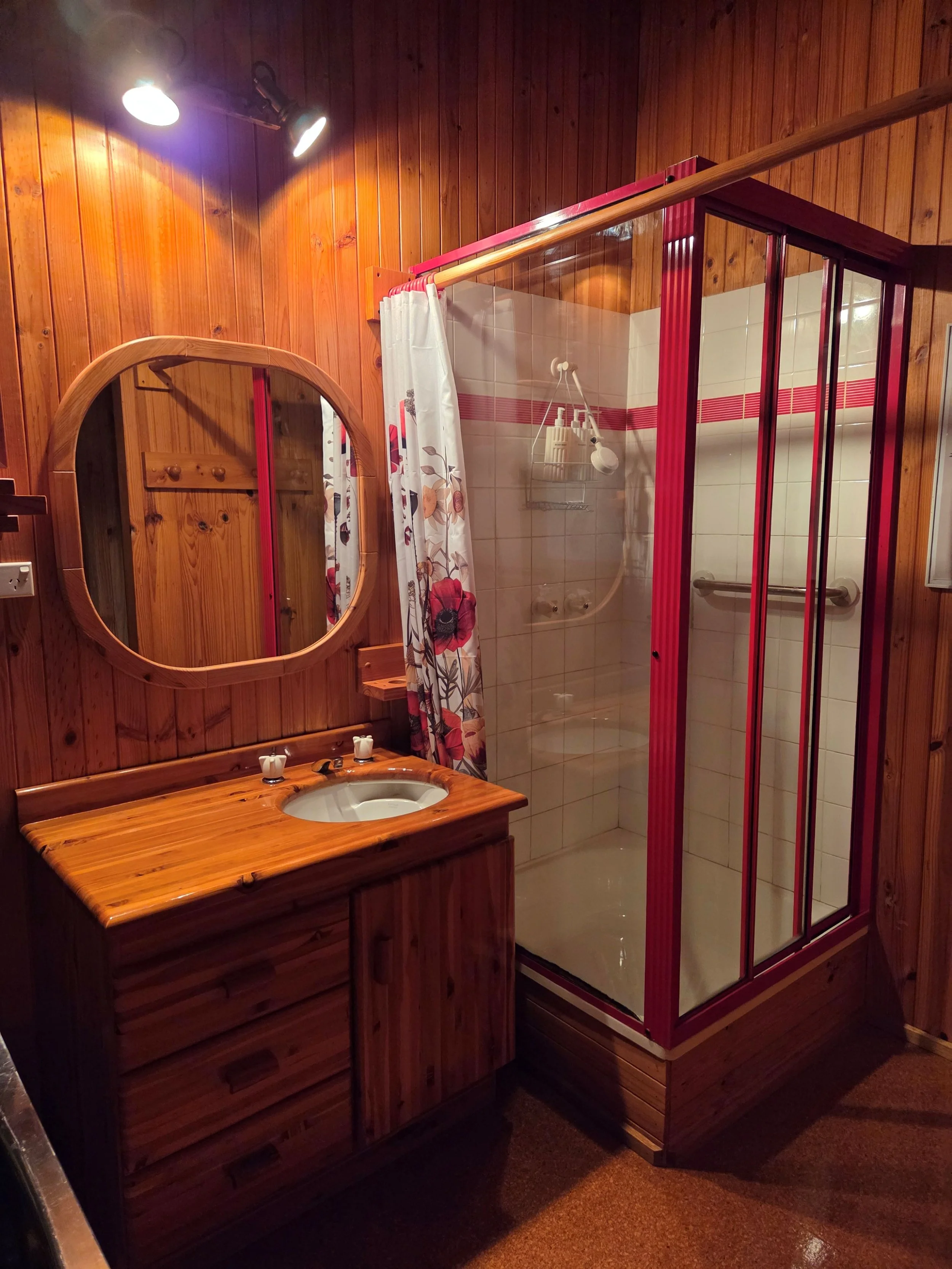 A bathroom with wooden walls and a red-framed glass shower stall. There is a wooden vanity with a sink, a mirror, and a floral shower curtain.