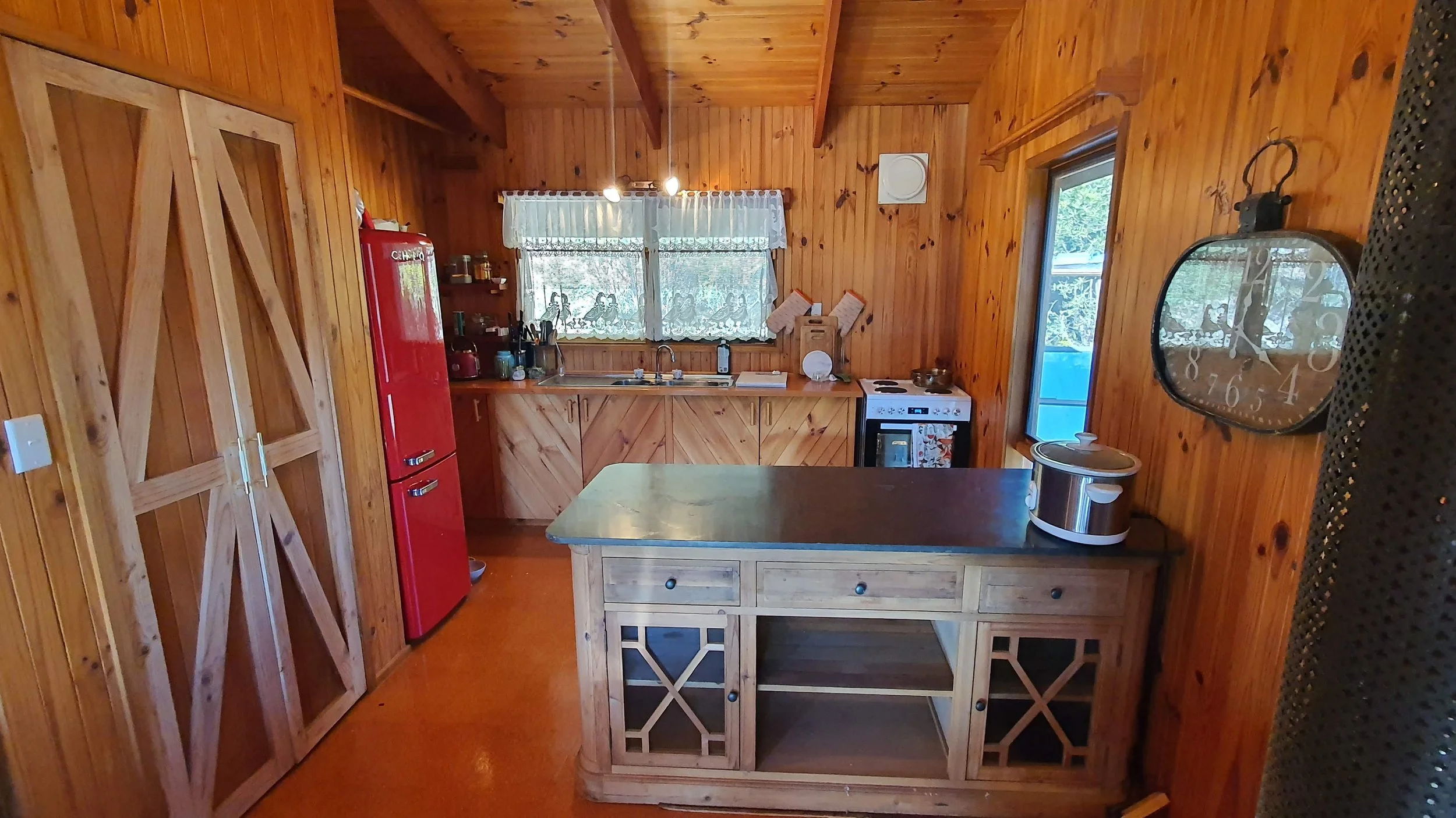 A cozy kitchen with wooden walls, ceiling, and cabinetry. There is a red refrigerator, a small stove, a sink under two windows with lace curtains, and various kitchen items on the counter. A wooden island with drawers is in the center. A large, decor