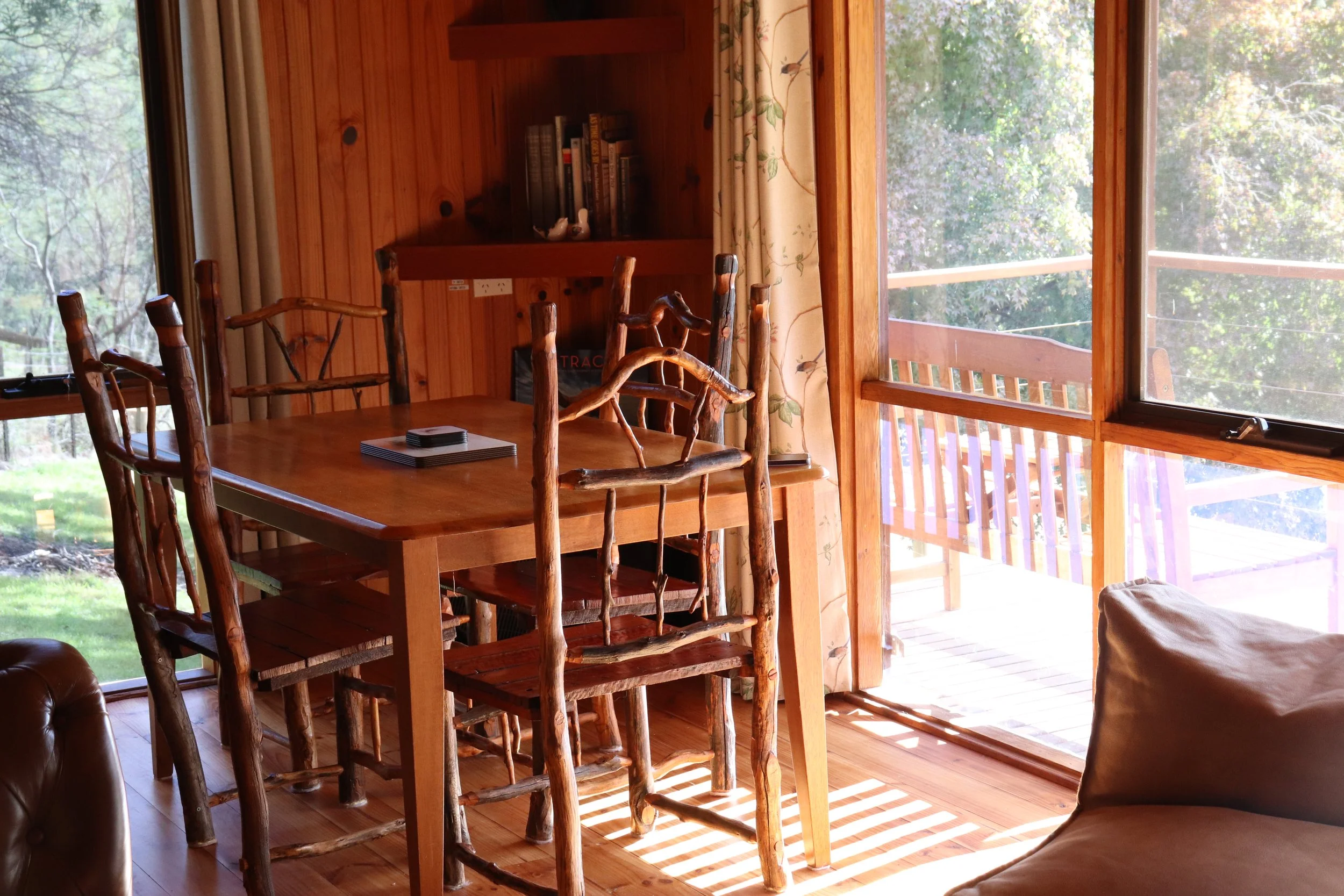 Interior of a wooden cabin or house with a dining table and rustic chairs, large glass windows showing trees outside, and a bookshelf on the wall.