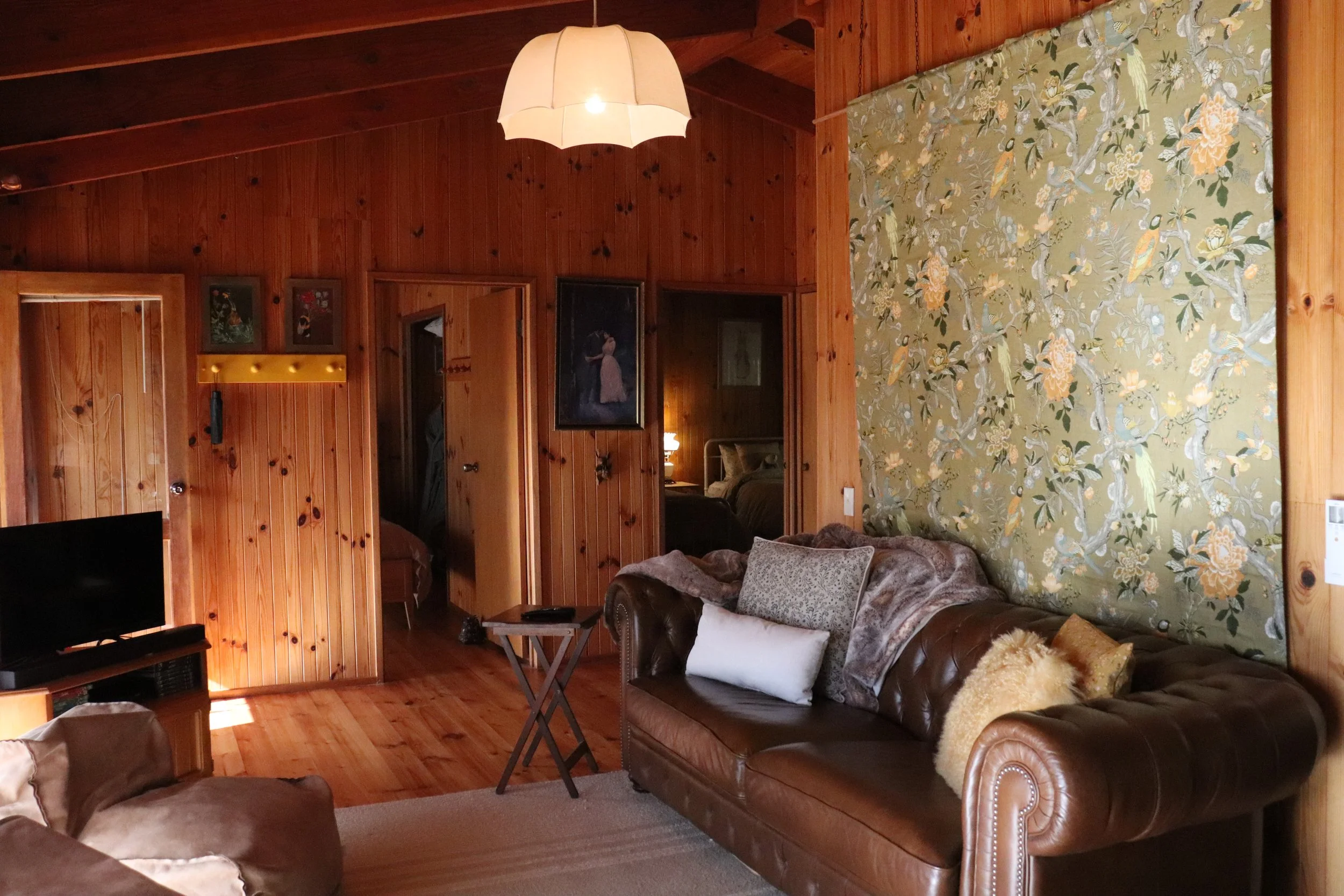 A cozy living room with wooden paneled walls, a leather sofa with pillows, a floral wall hanging, and a pendant lamp hanging from the ceiling.