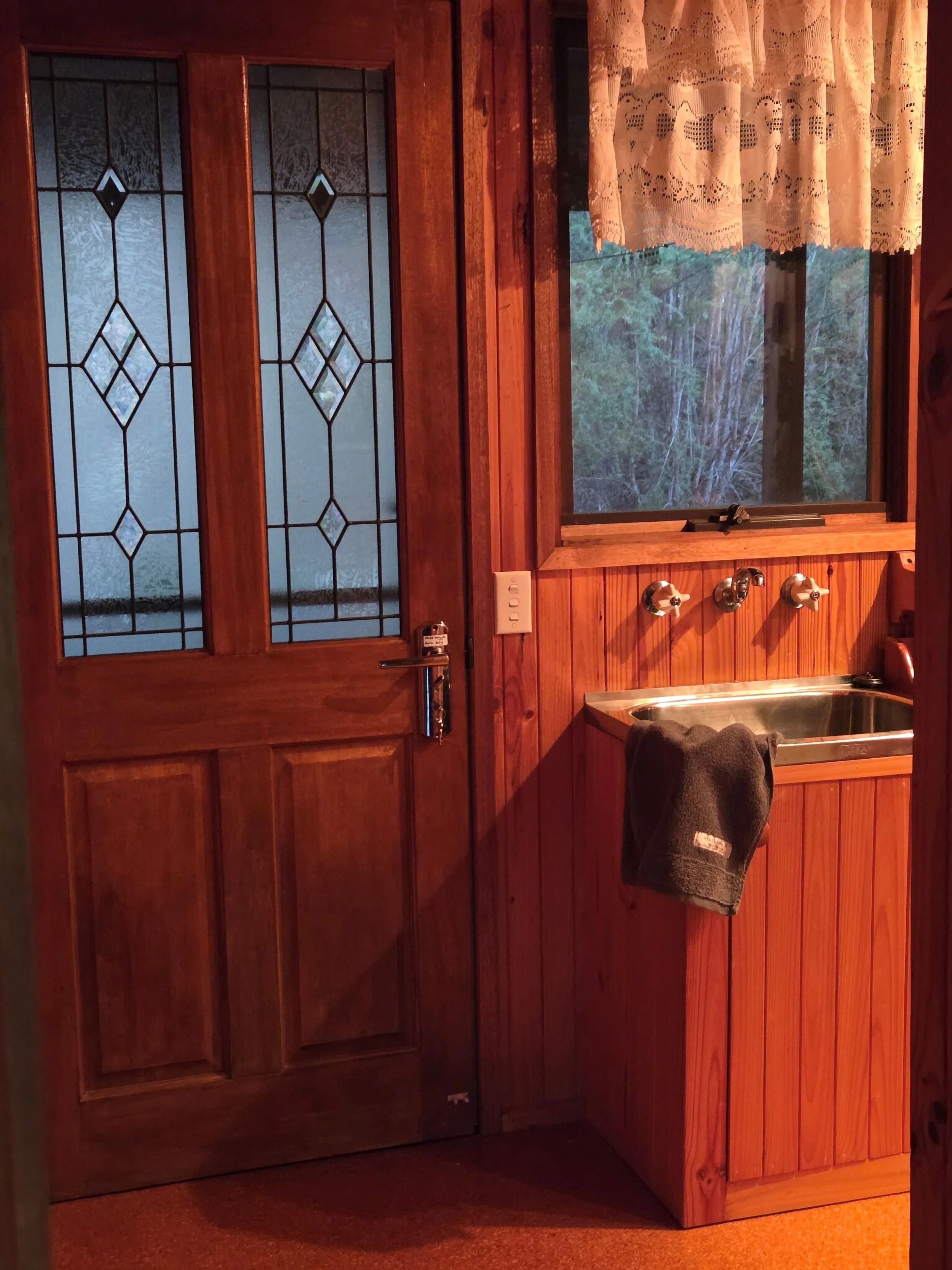 A wooden kitchen counter with a stainless steel sink, a gray towel hanging on the front, and three silver faucets mounted on a wooden panel. A small window with lace curtains and a view of green trees outside, situated next to a wooden door with deco