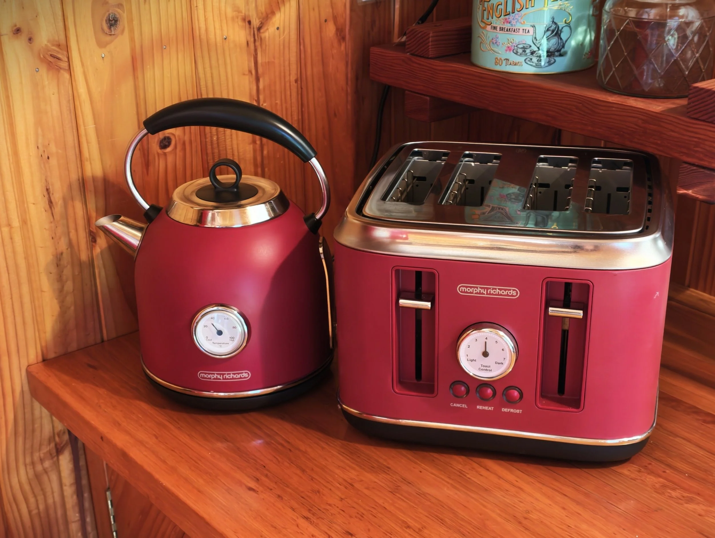 Retro look kettle and pink toaster on a wooden countertop against a wooden wall, with a decorative tin and container on a shelf above.
