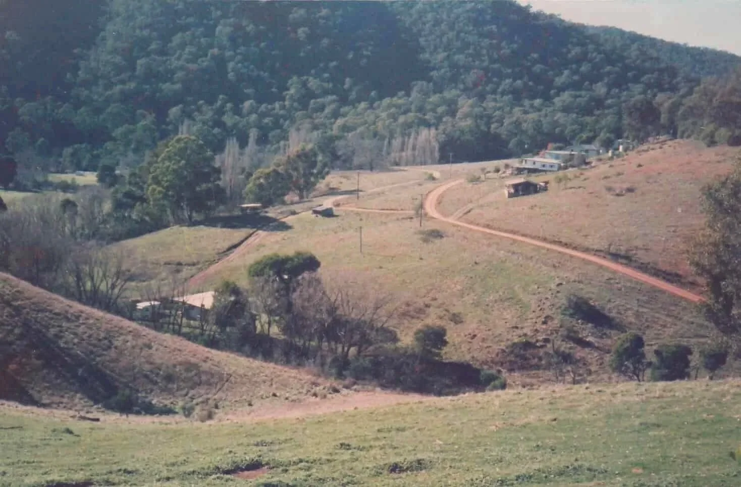 A rural landscape with rolling hills, a few scattered houses, dirt roads, and a backdrop of forested mountains.