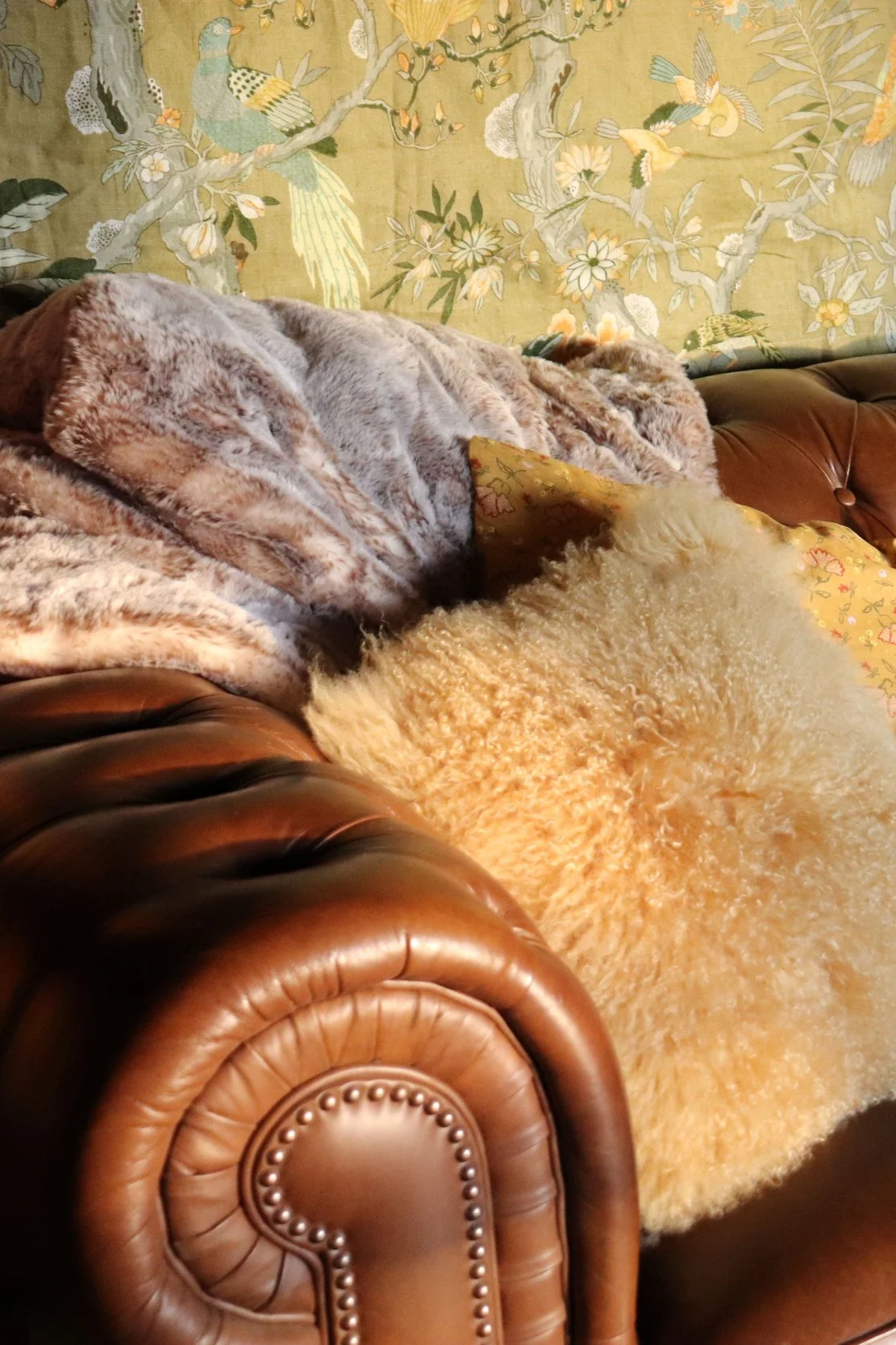 A brown leather couch with a beige fluffy blanket and a yellow floral pillow, against a wallpapered background with birds and branches.