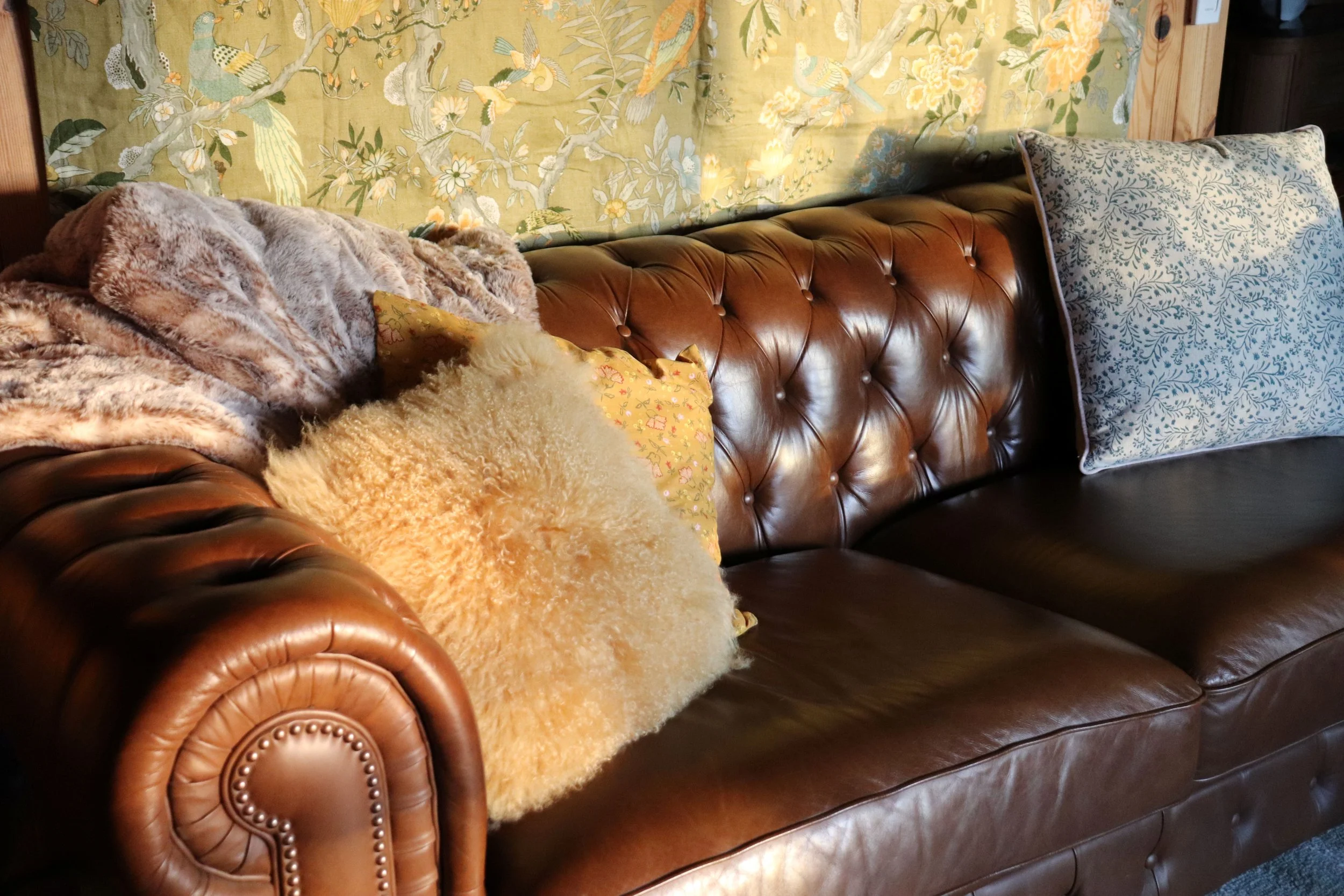 A brown leather tufted sofa with pillows, a plush blanket, and a vintage floral wallpaper background.