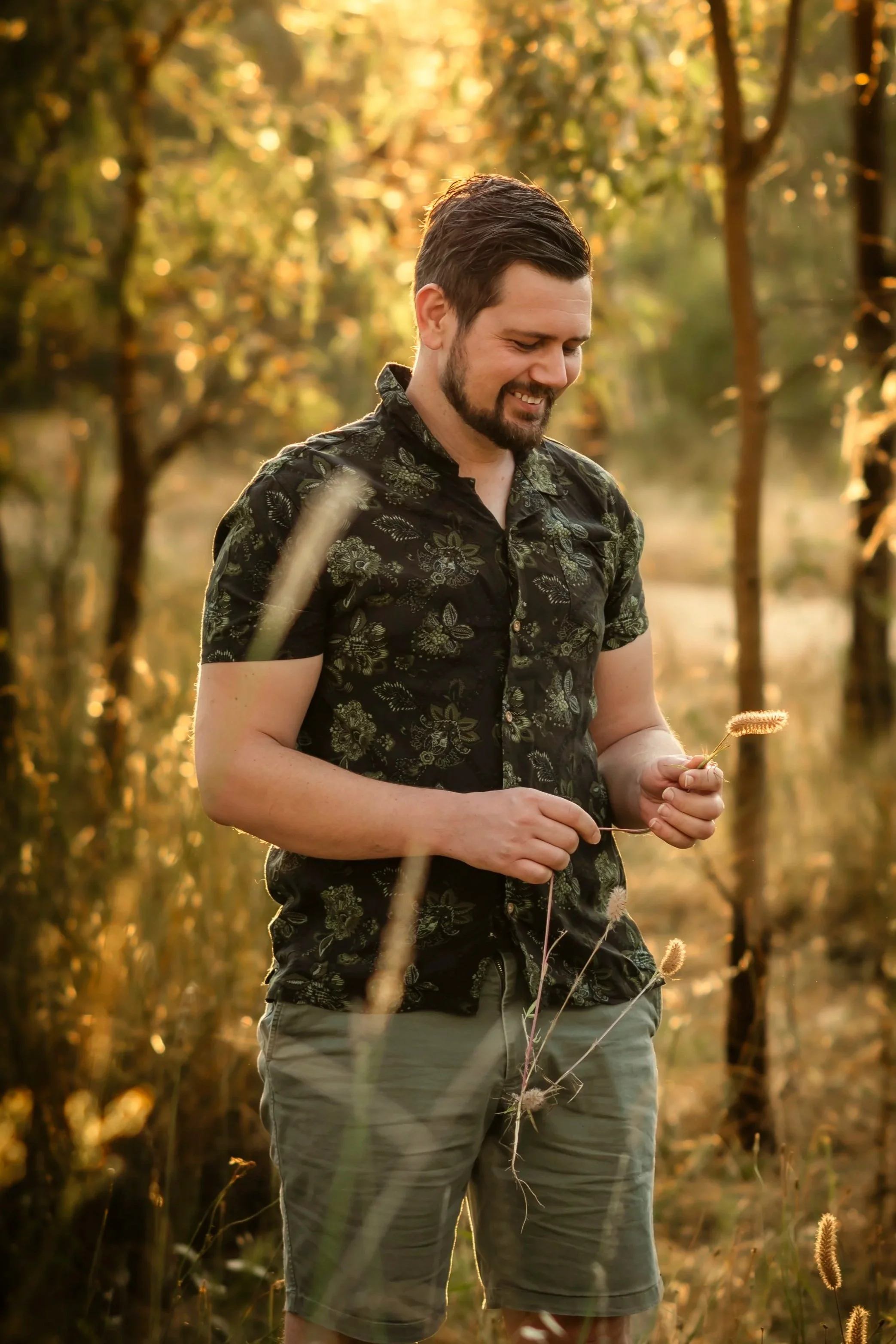 A man in a black floral shirt and cargo shorts smiling and holding a plant in a sunlit forest during golden hour.