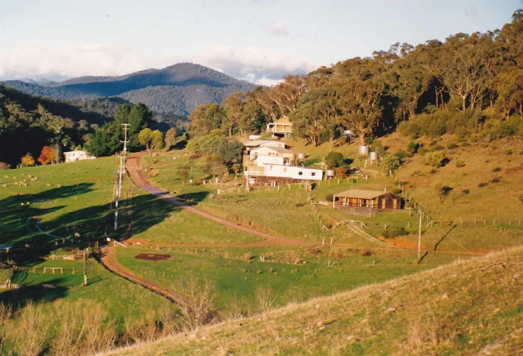 A rural landscape with rolling green hills, scattered houses and farm buildings, and trees on a hillside, with mountains in the background and a partly cloudy sky.