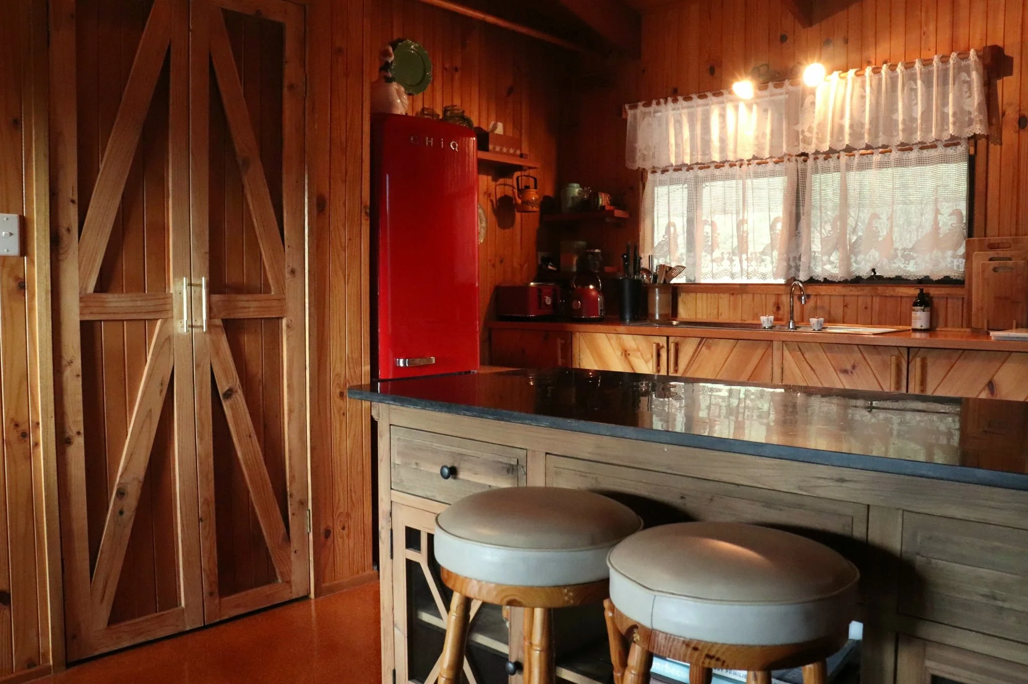 Kitchen with wooden panel walls, red vintage fridge, small wooden kitchen island or table, two round wooden stools, and a window with lace curtains.