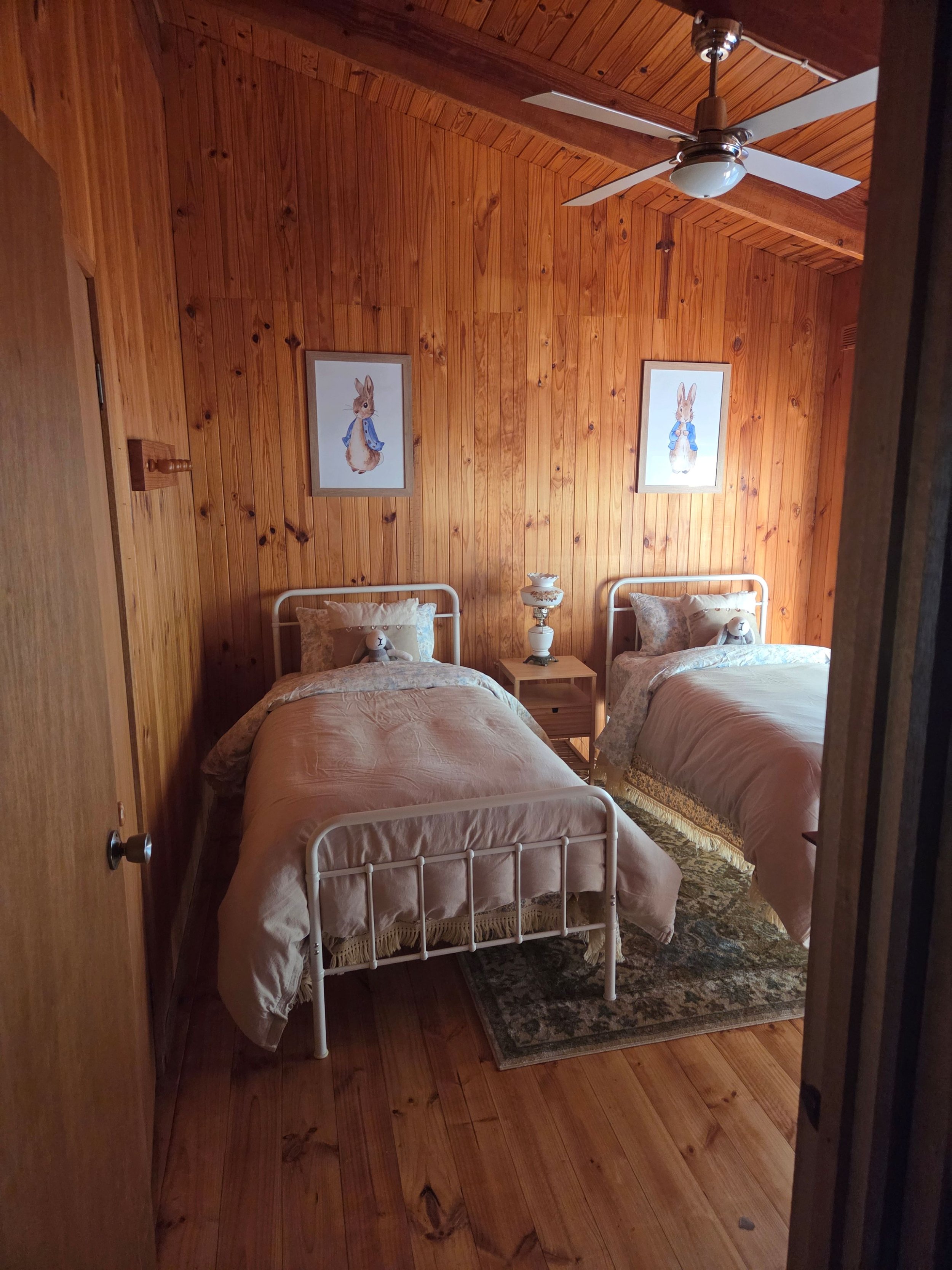 A bedroom with wooden paneling on the walls and ceiling, featuring two twin beds with beige bedspreads, a nightstand with a vintage lamp, and framed pictures of rabbits dressed in blue shirts hanging on the wall. There is a ceiling fan with a light f