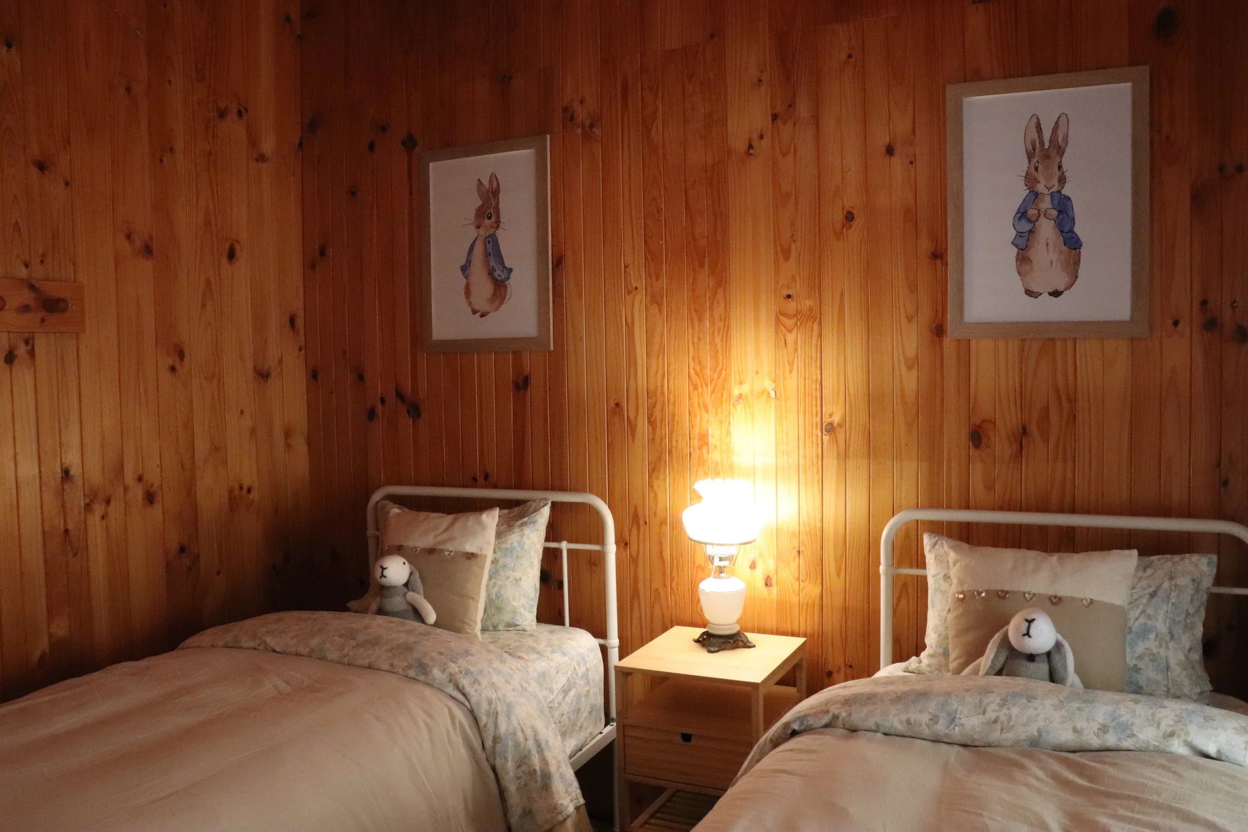 A bedroom with wooden paneled walls, two twin beds with floral bedspreads, stuffed bunny toys, a nightstand with a lamp, and framed pictures of bunnies in blue jackets on the wall.