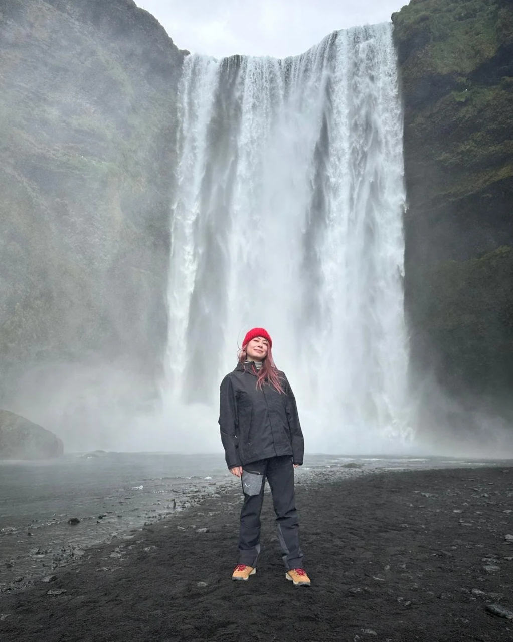 A woman stands in front of a tall waterfall, wearing a black jacket, gray pants, red boots, and a red beanie, smiling at the camera.
