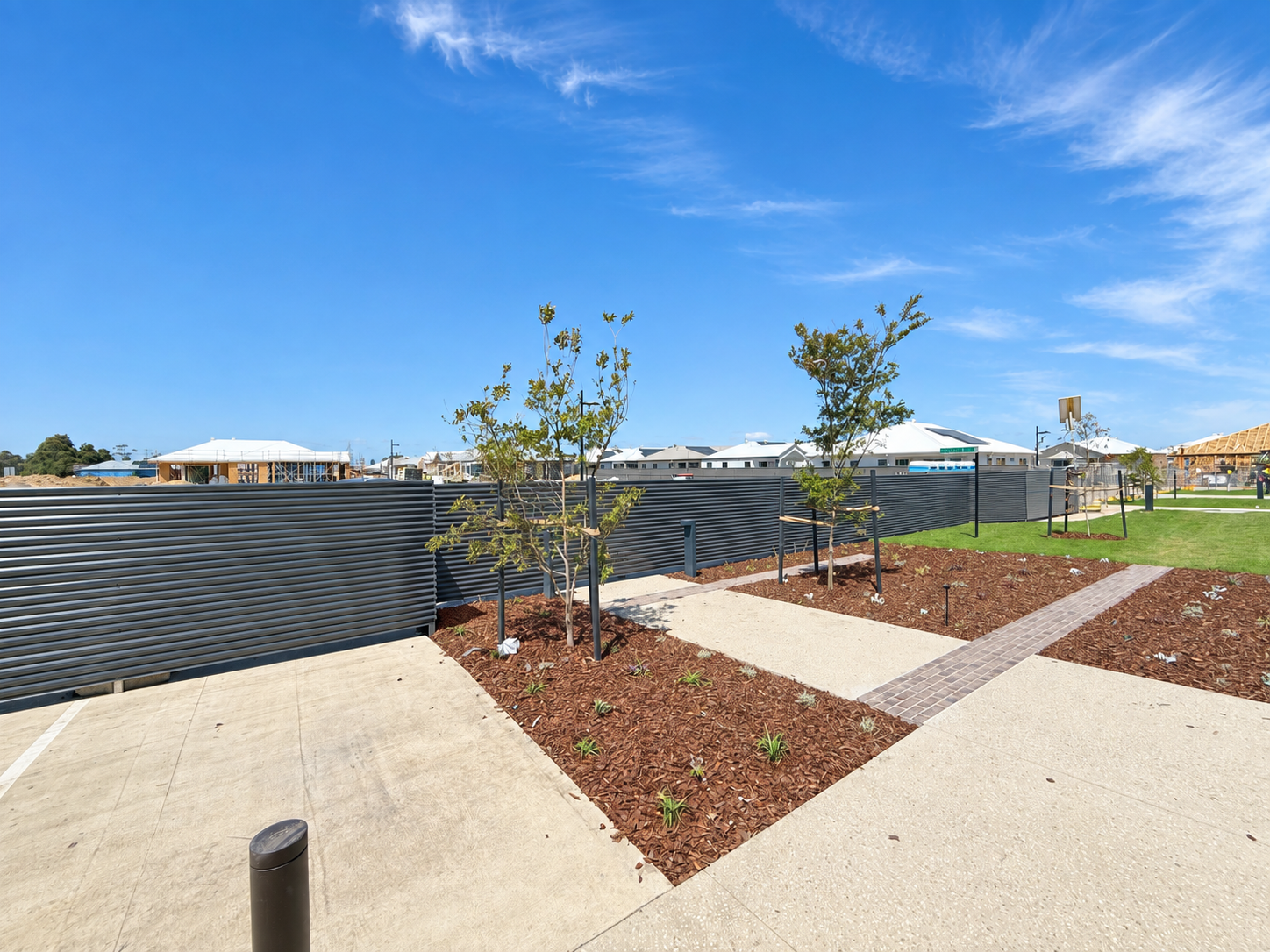 A sunny day outdoor scene with a sidewalk, small trees, and a black metal fence in front of stay-under-construction buildings and clear blue sky.