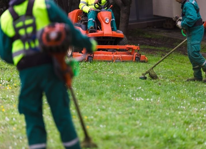 People maintaining a grassy area with gardening tools, and a person operating an orange riding lawn mower.