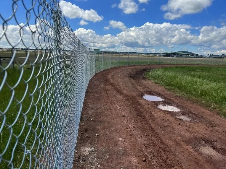 A dirt baseball or softball field with a chain-link fence on the left and a grassy area on the right under a partly cloudy sky.