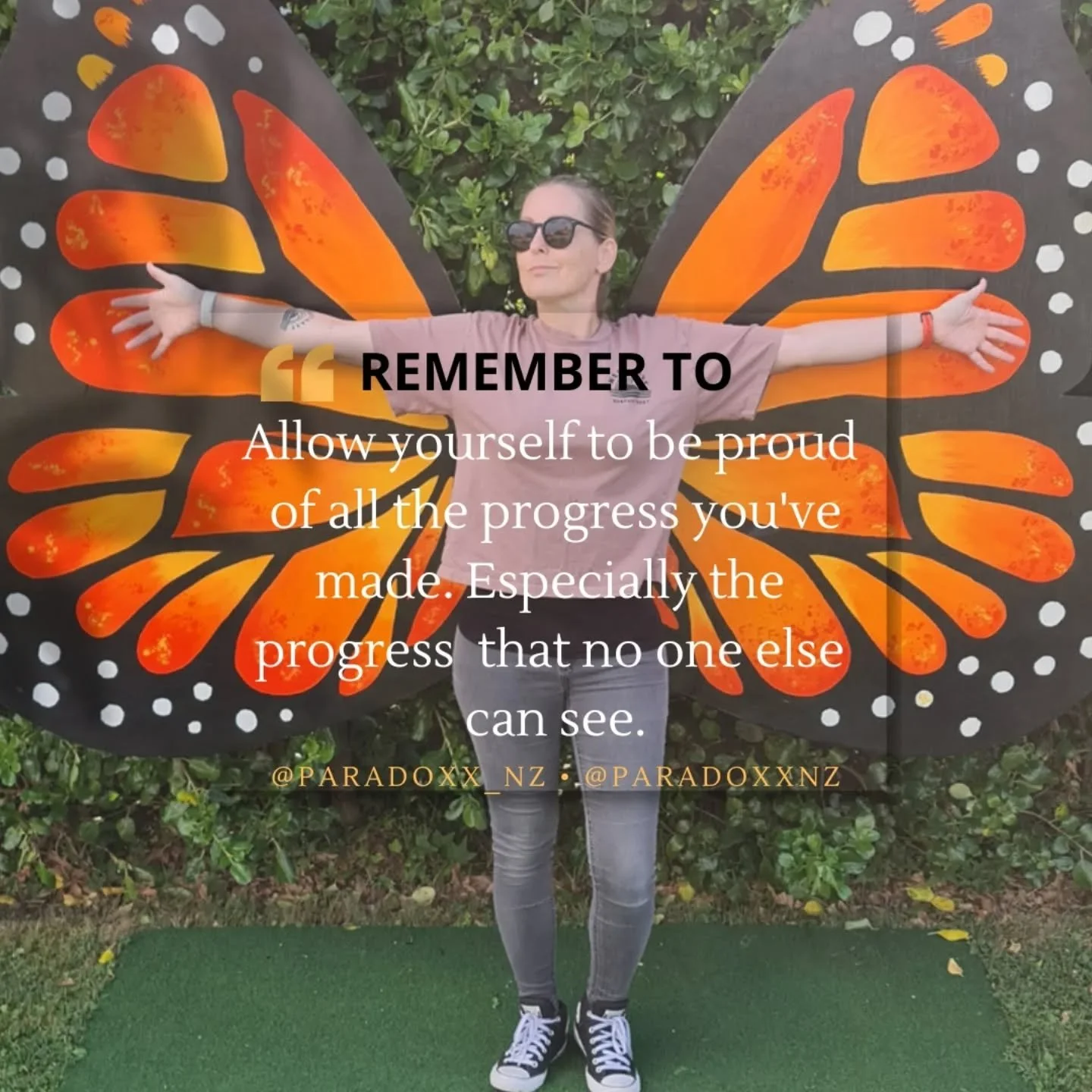 Kath - Nurse Therapist stands with arms outstretched in front of butterfly wings painting, wearing a light pink T-shirt, gray jeans, and black Converse sneakers. The background features green bushes and grass.