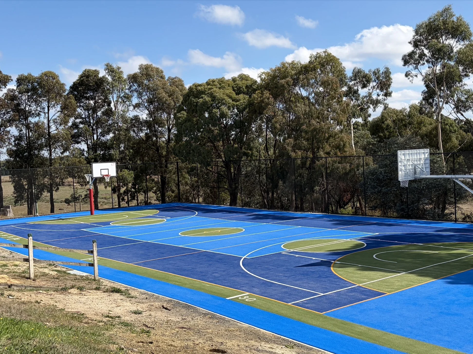 Outdoor multi sport court with colorful surface and two basketball hoops, surrounded by a black chain-link fence and tall trees under a partly cloudy sky.