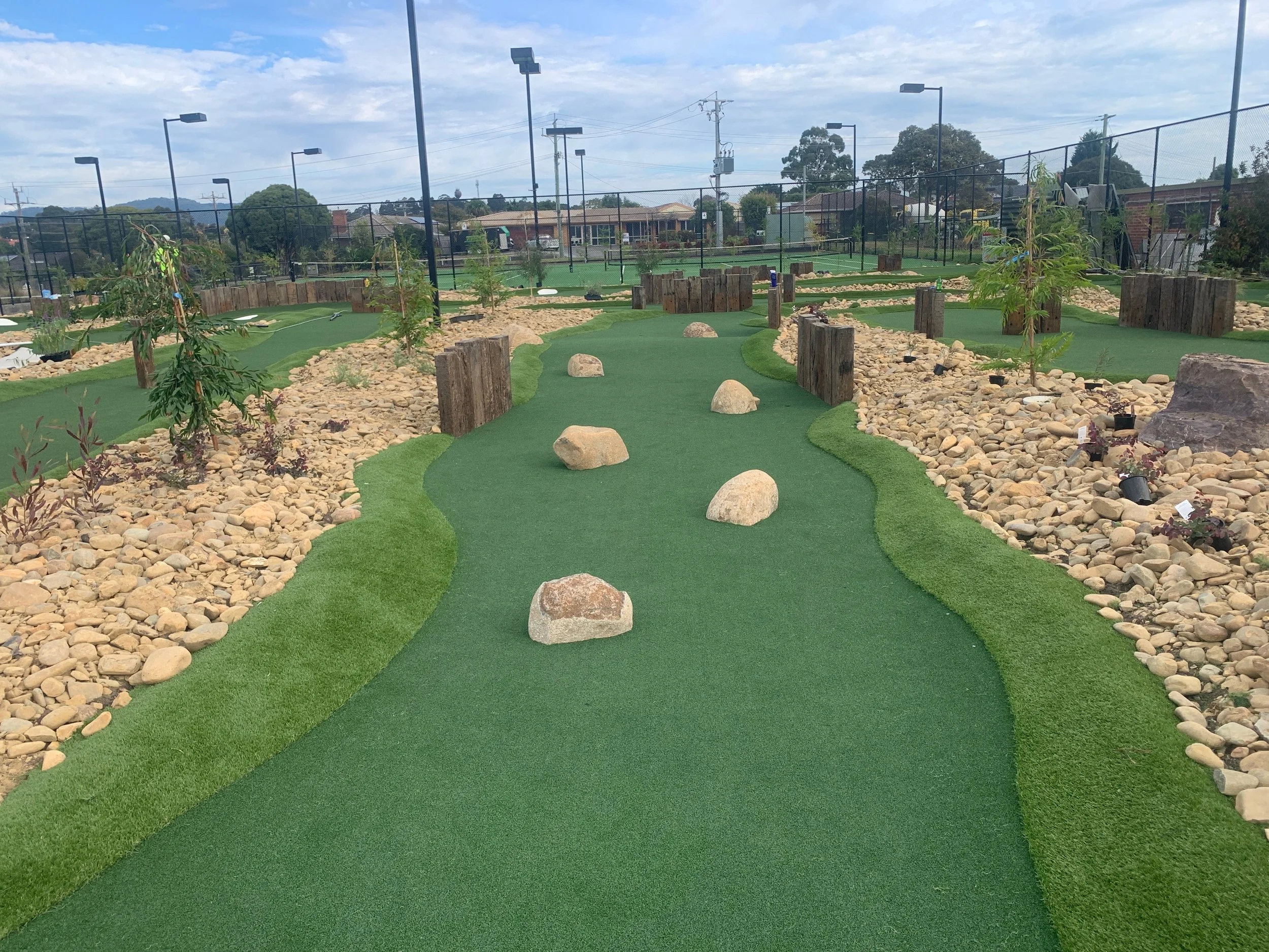 An astro turf mini golf course with rocks, small trees, wooden barriers, and black lighting fixtures, surrounded by a chain-link fence and residential neighborhood in the background.