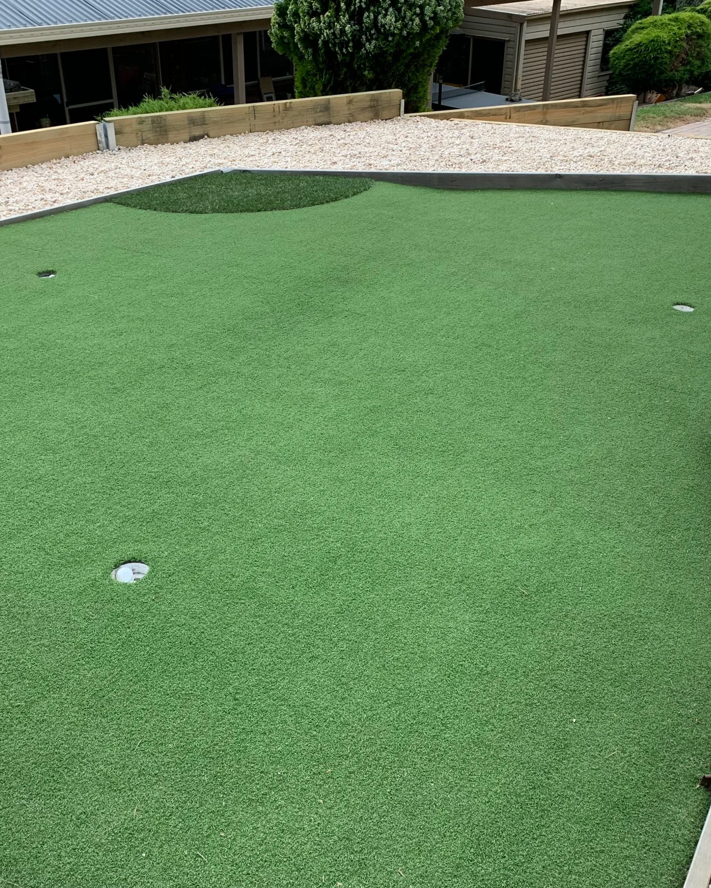 Artificial putting green with golf holes, surrounded by a white rock filled perimeter.  wooden retaining wall, and residential buildings and trees in the background.