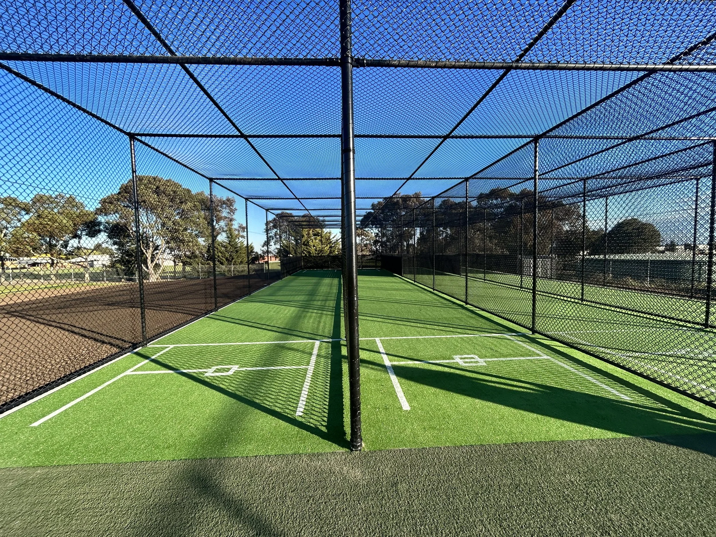 A synthetic cricket wicket enclosed by a tall chain-link fence under a blue sky with trees in the background.