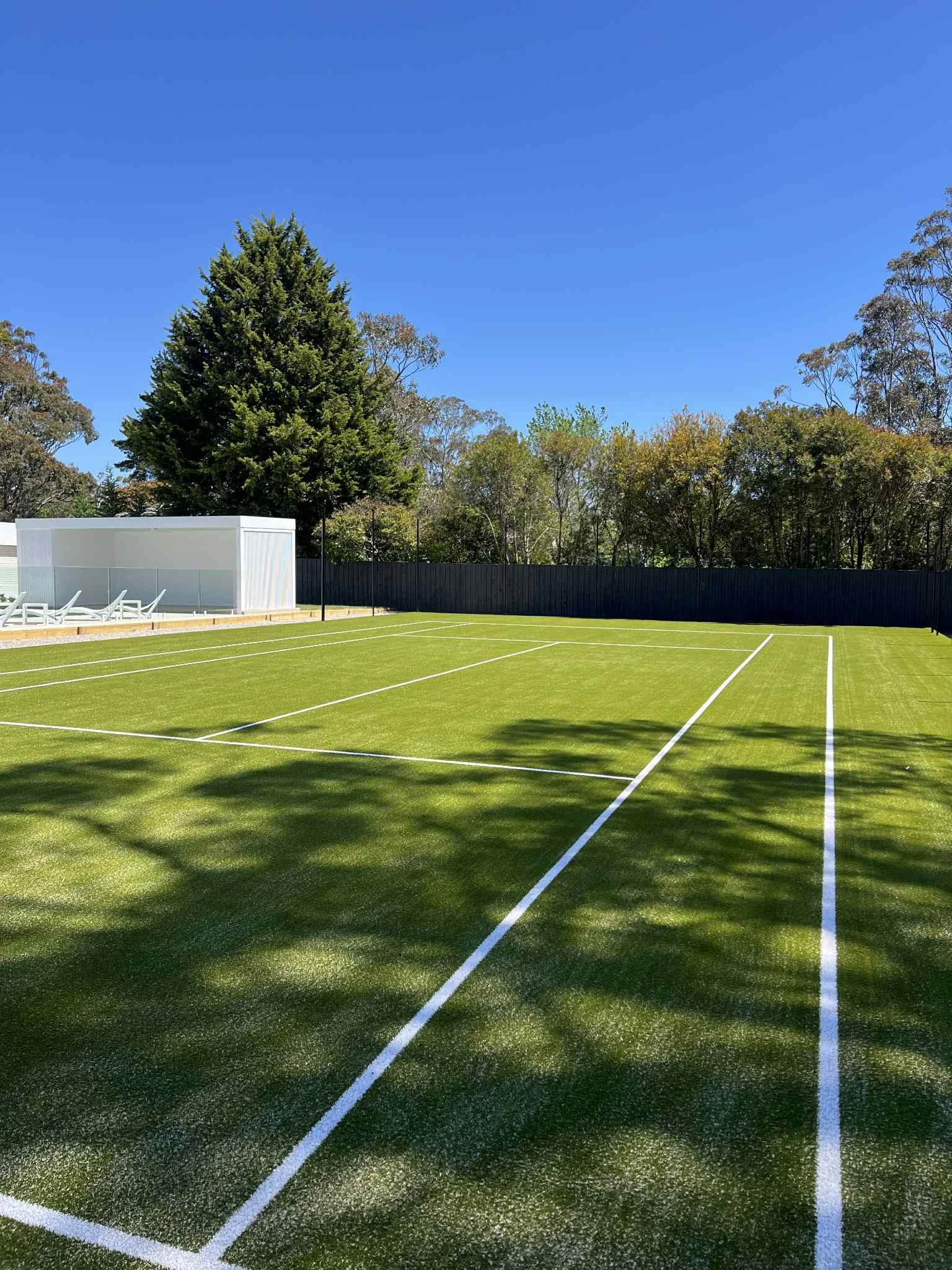 synthetic grass tennis court with bright green surface, white lines, and a backdrop of trees and a blue sky.