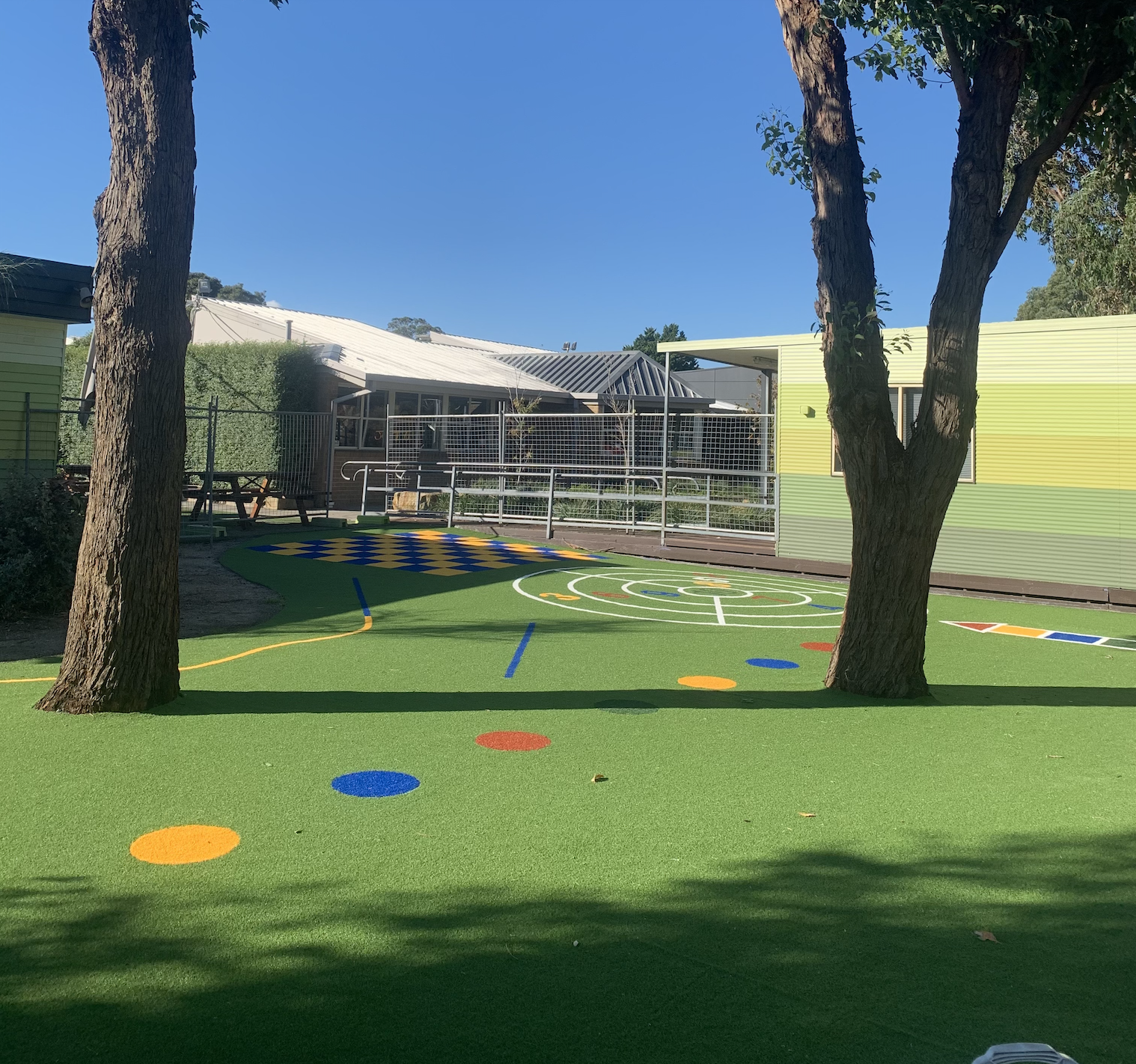 Colourful synthetic grass play area at a primary school, trees, and a building in the background.