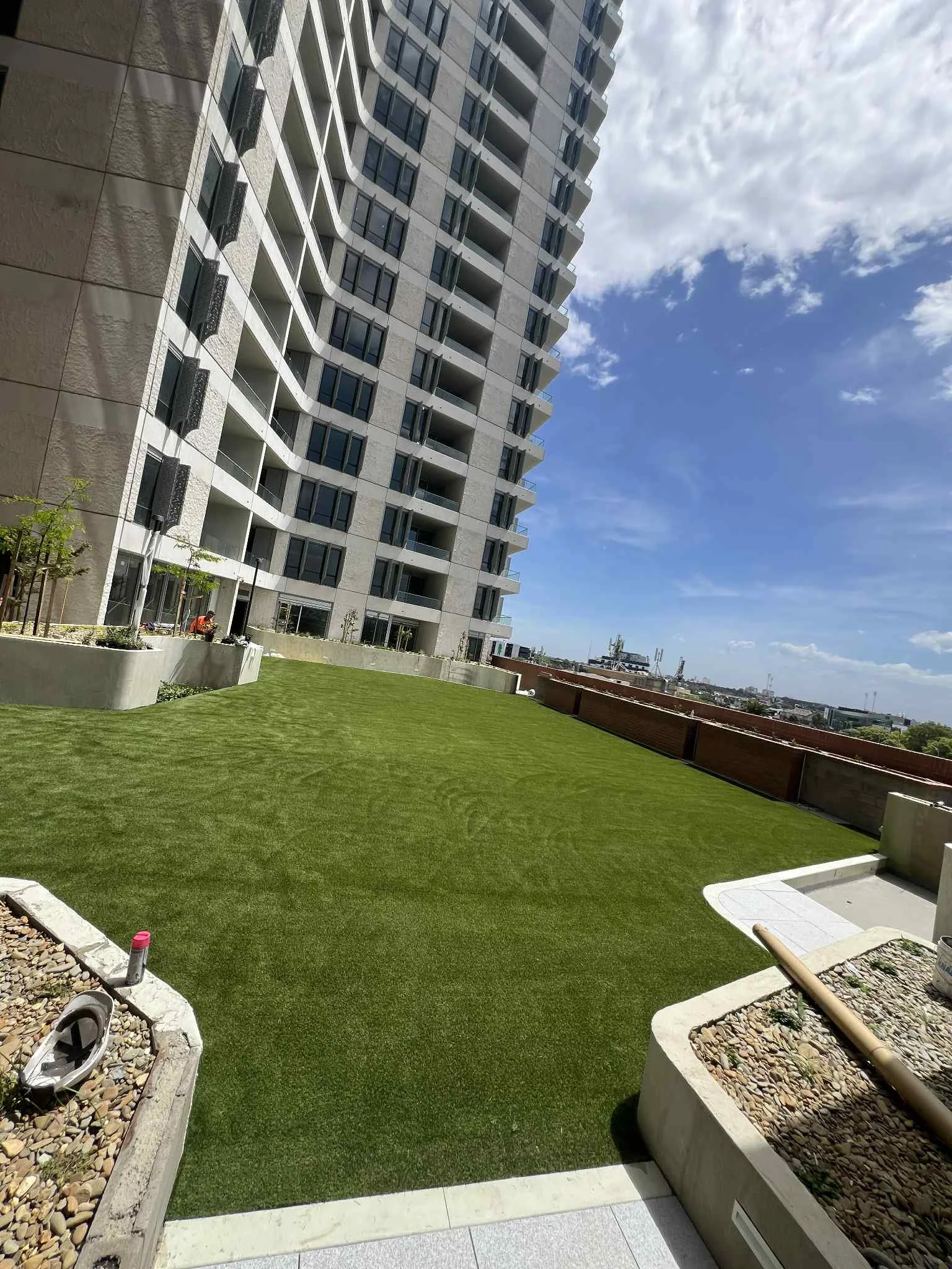 View of a modern high-rise apartment building with a landscaped artificial turf in the foreground, under a partly cloudy sky.