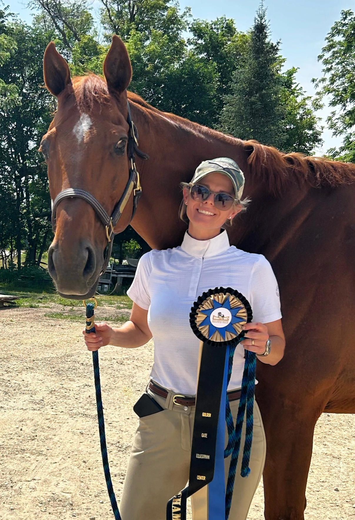 A woman smiling, wearing sunglasses and a white shirt, standing outdoors with a large brown horse. She holds a blue and black ribbon, and the horse is wearing a halter. The background has trees and a clear sky.